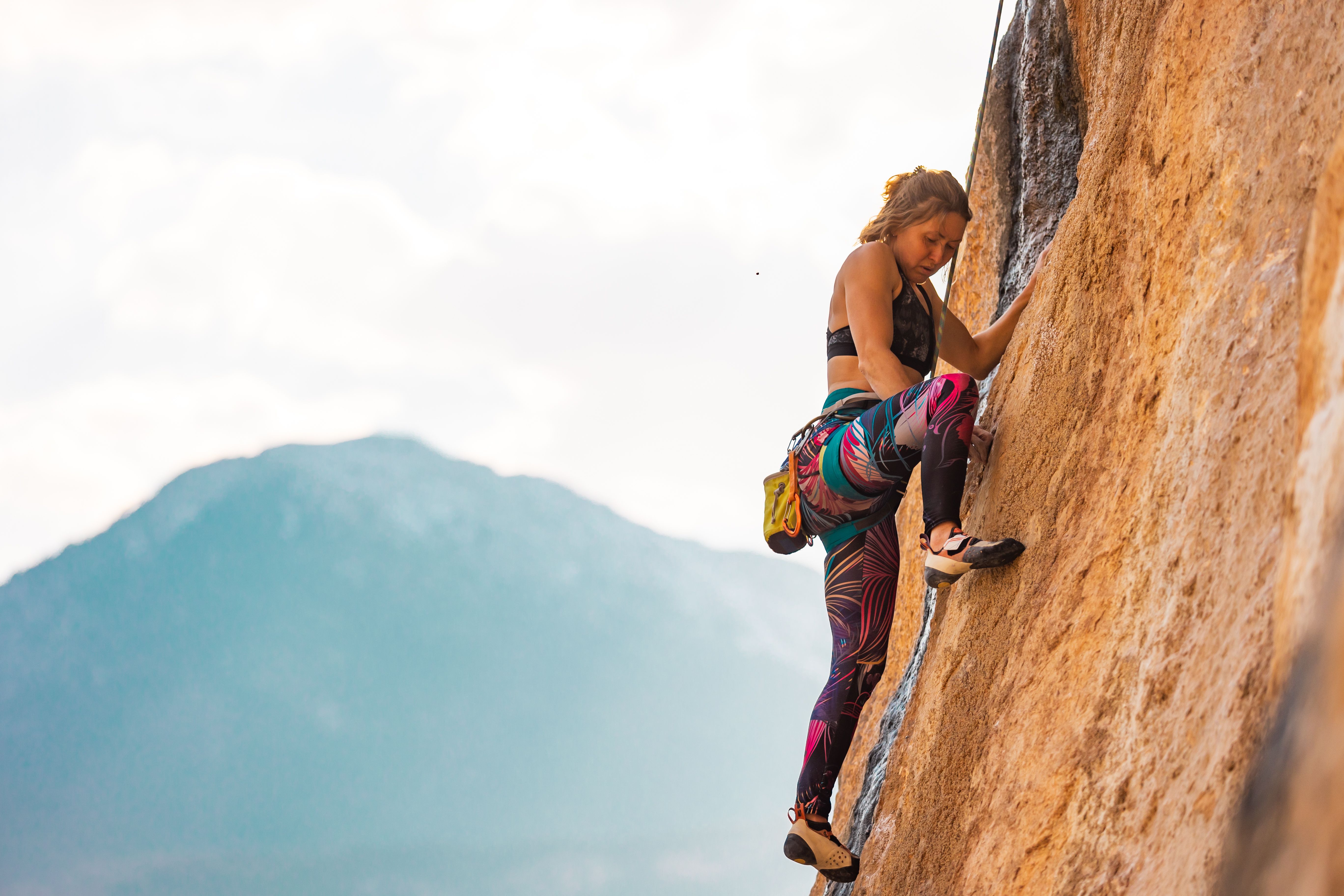 Woman climbing a vertical rock face with a mountain landscape in the background, wearing colorful leggings and rock-climbing gear