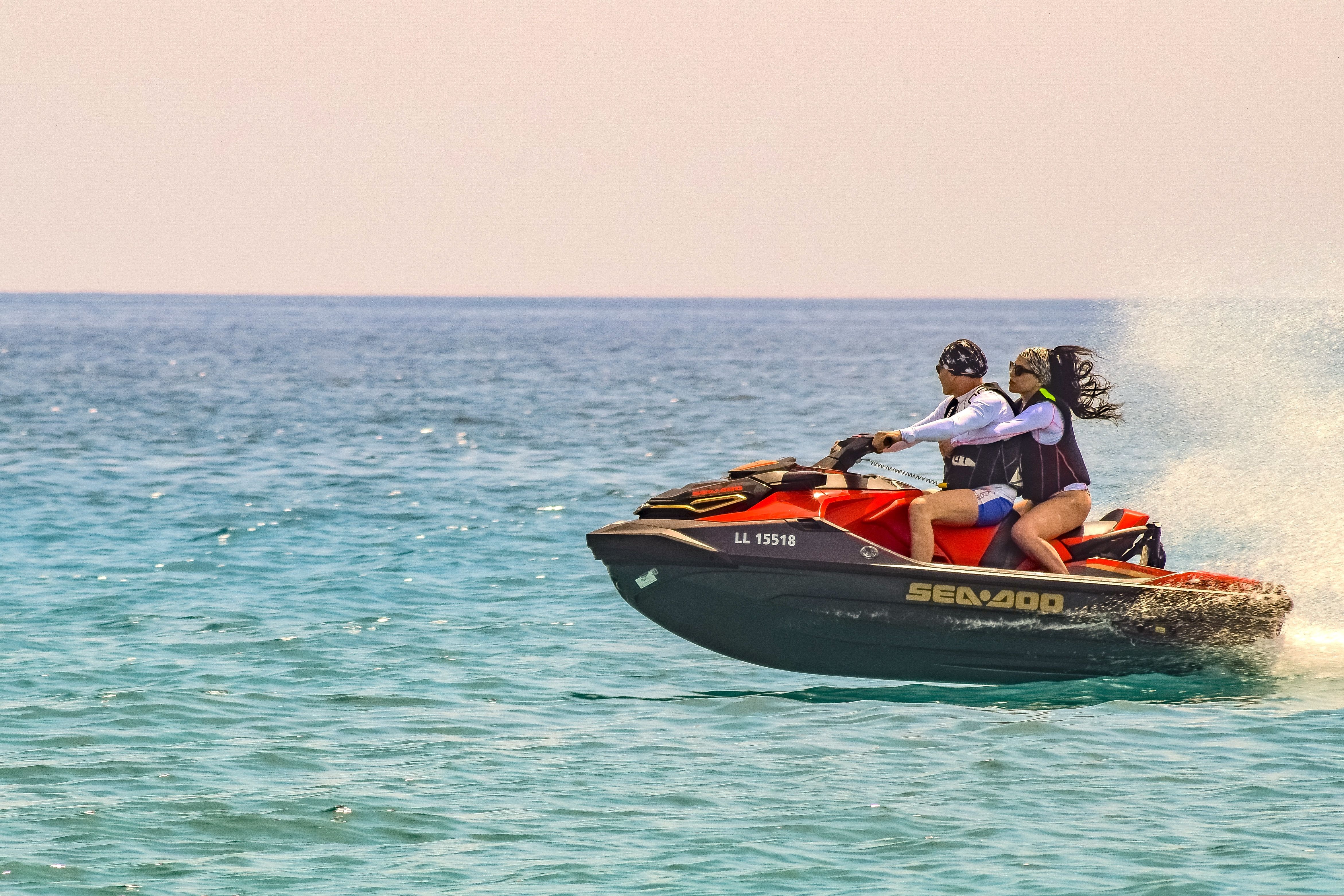 Two people riding a red jet ski across the ocean on a bright sunny day, leaving a trail of water behind
