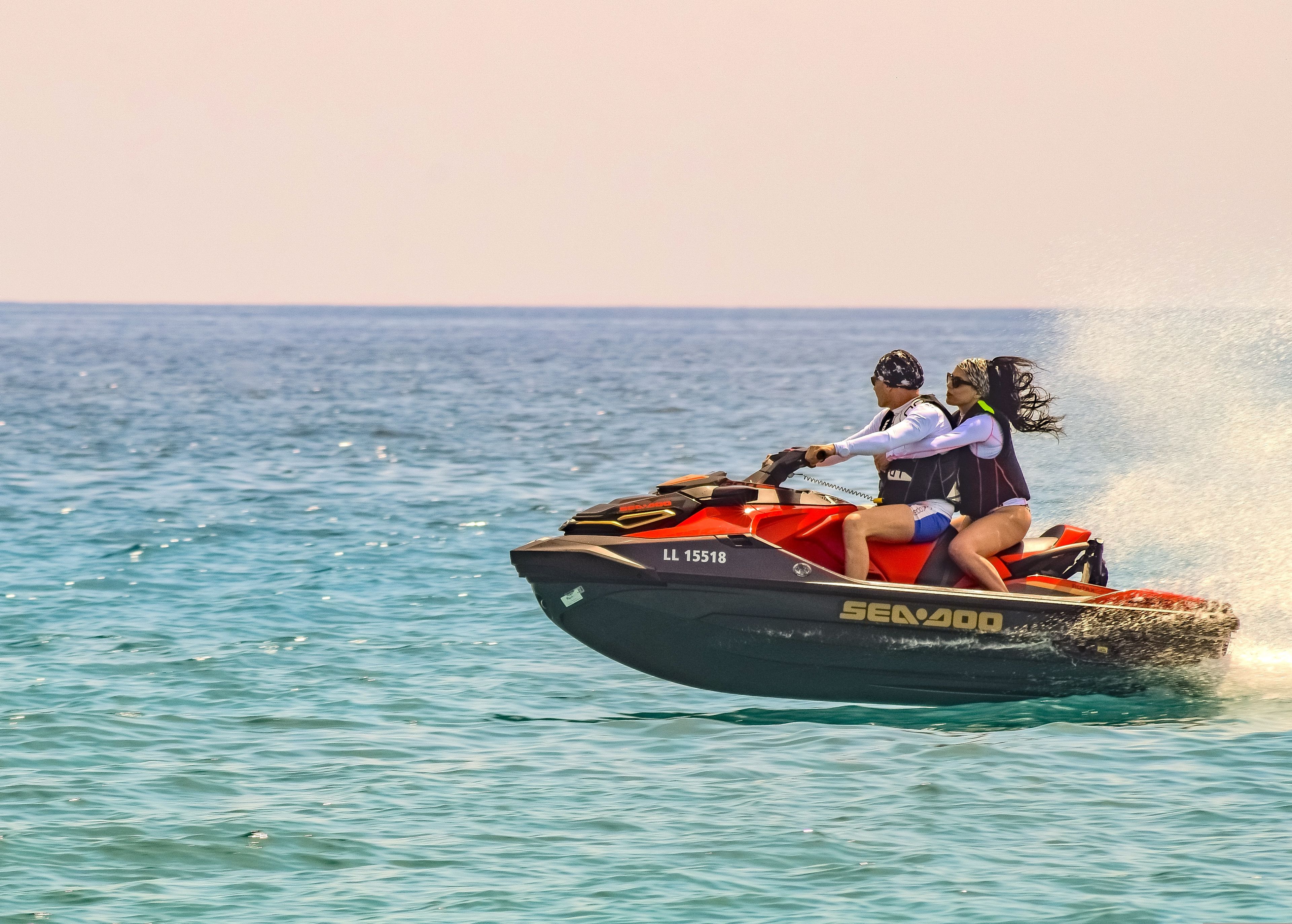 Two people riding a red jet ski across the ocean on a bright sunny day, leaving a trail of water behind
