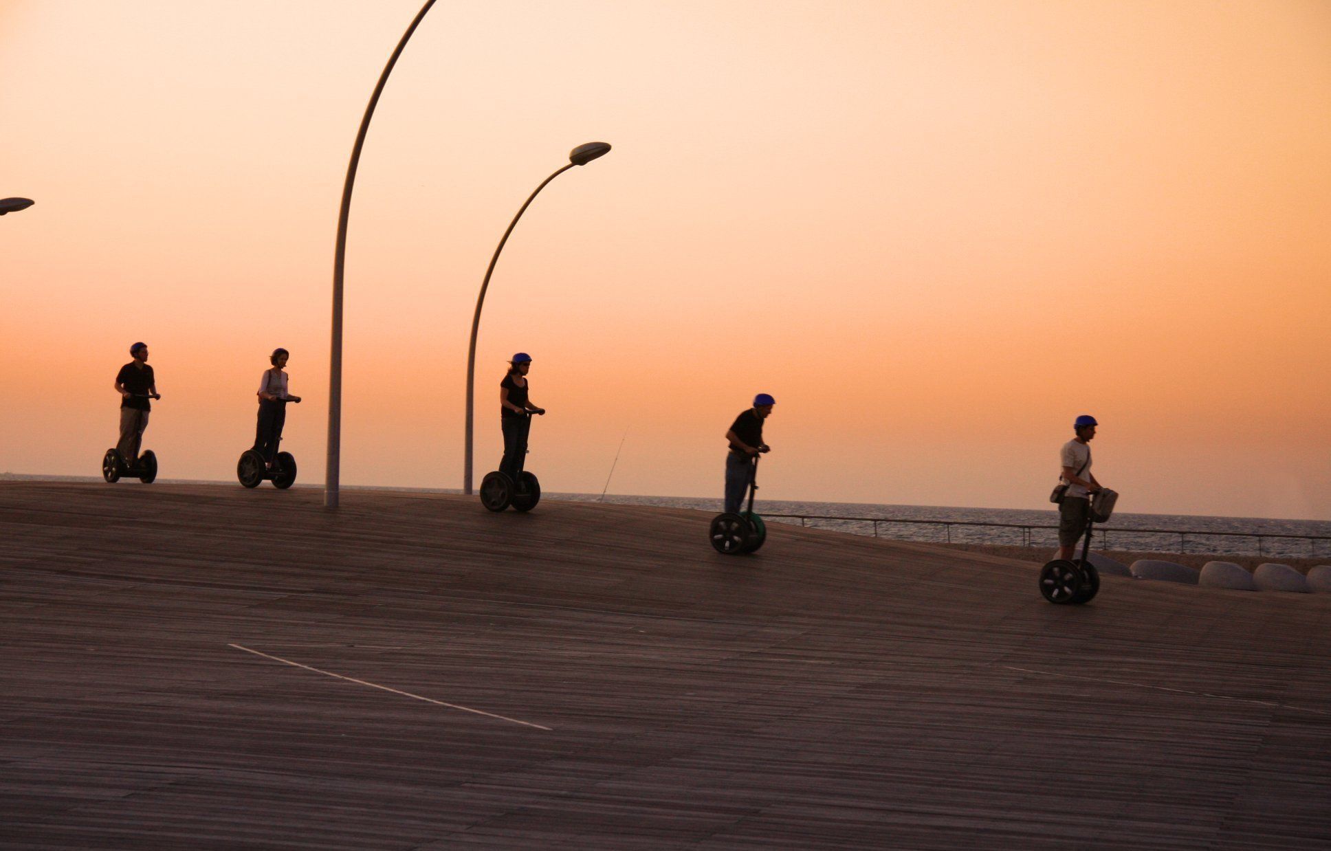 Group of people riding Segways along a boardwalk at sunset, with modern streetlights and the sea in the background