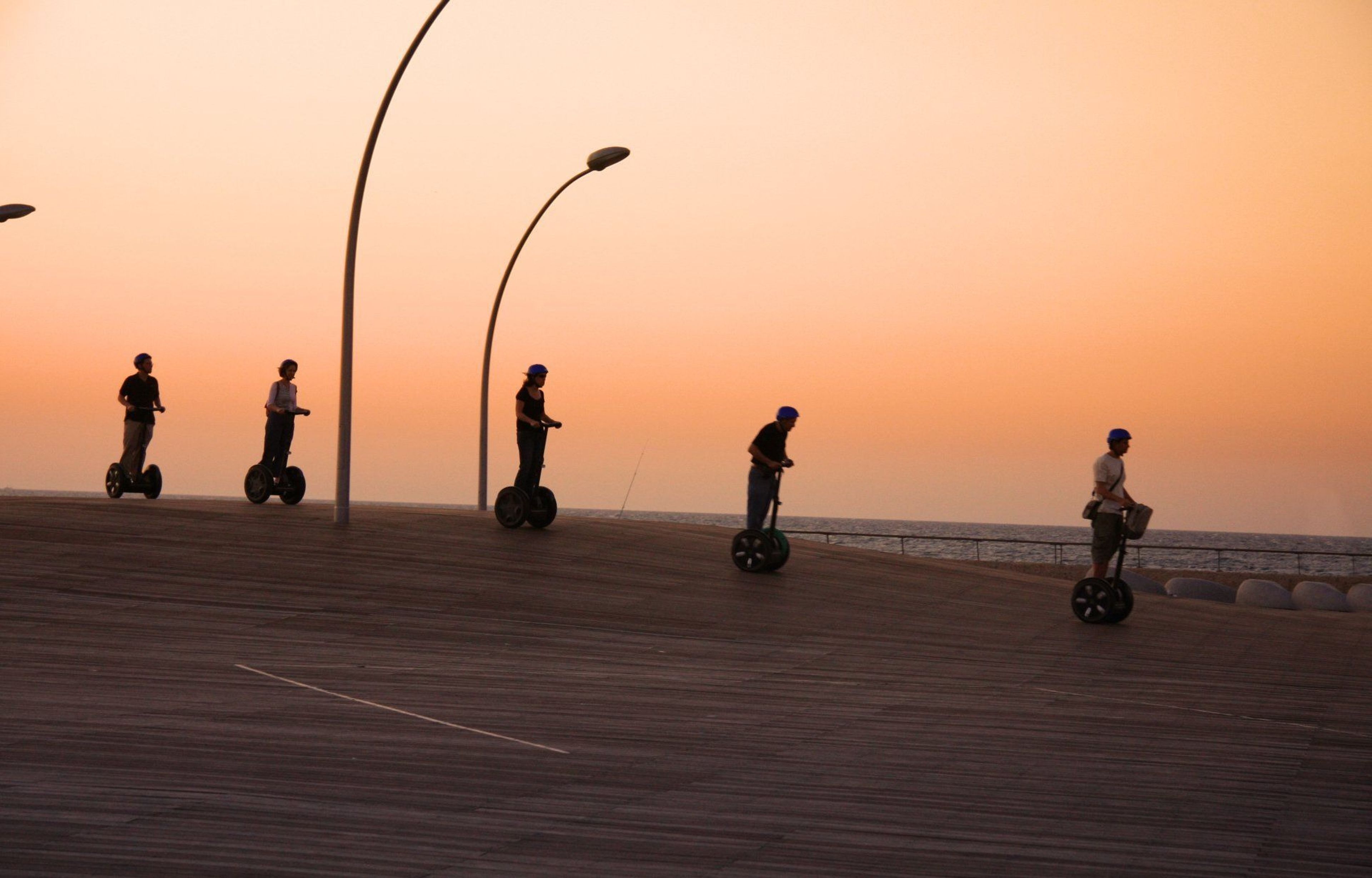 Group of people riding Segways along a boardwalk at sunset, with modern streetlights and the sea in the background