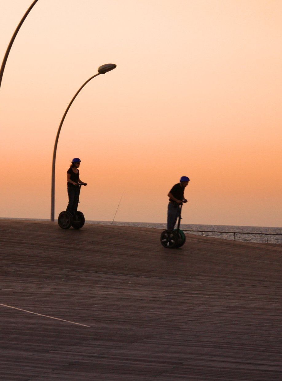 Group of people riding Segways along a boardwalk at sunset, with modern streetlights and the sea in the background