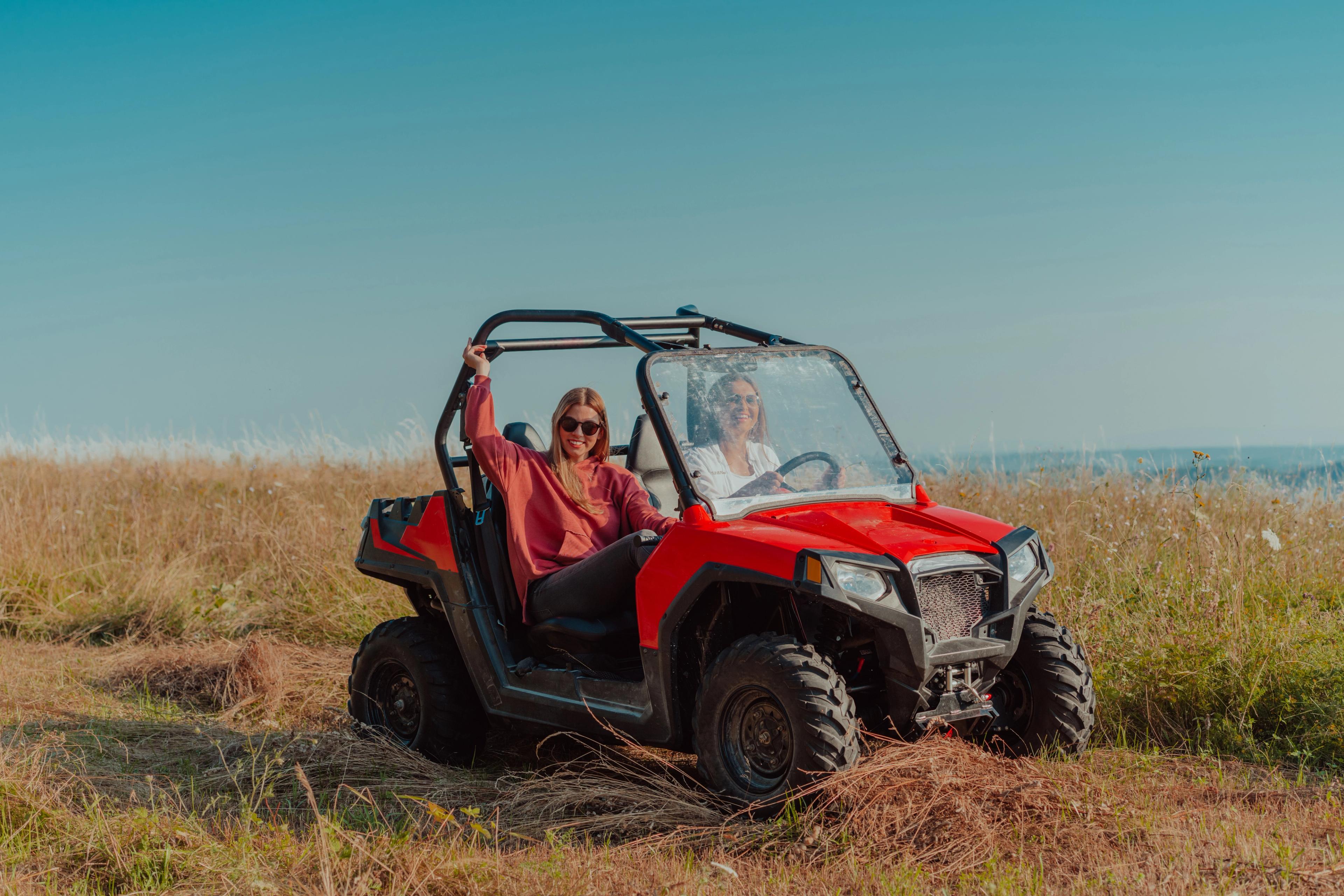Two women smiling and riding a buggy in an open grassy field under a clear sky