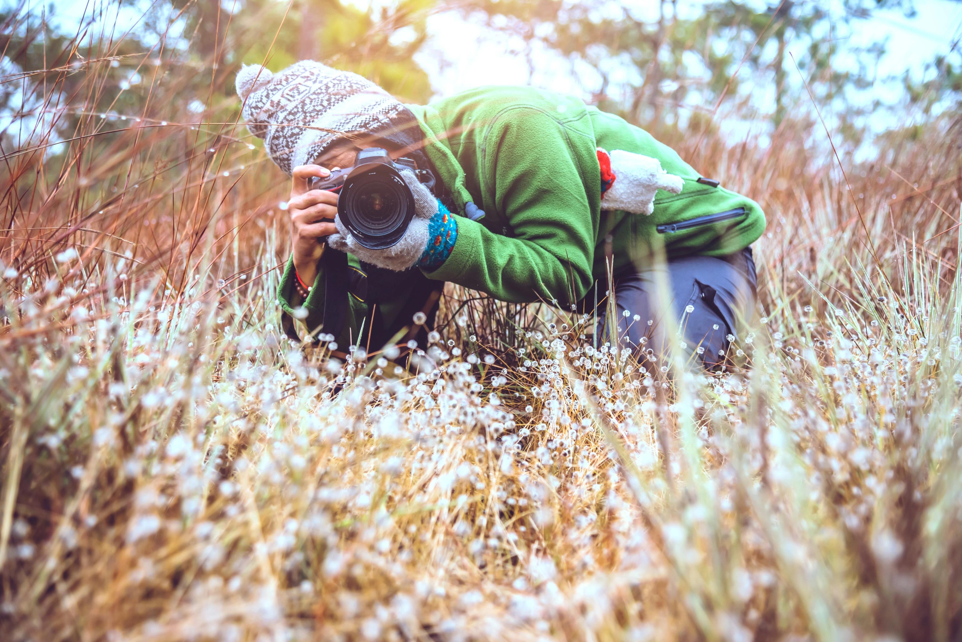 A photographer crouched low in a grassy field, aiming their camera to capture close-up shots of nature