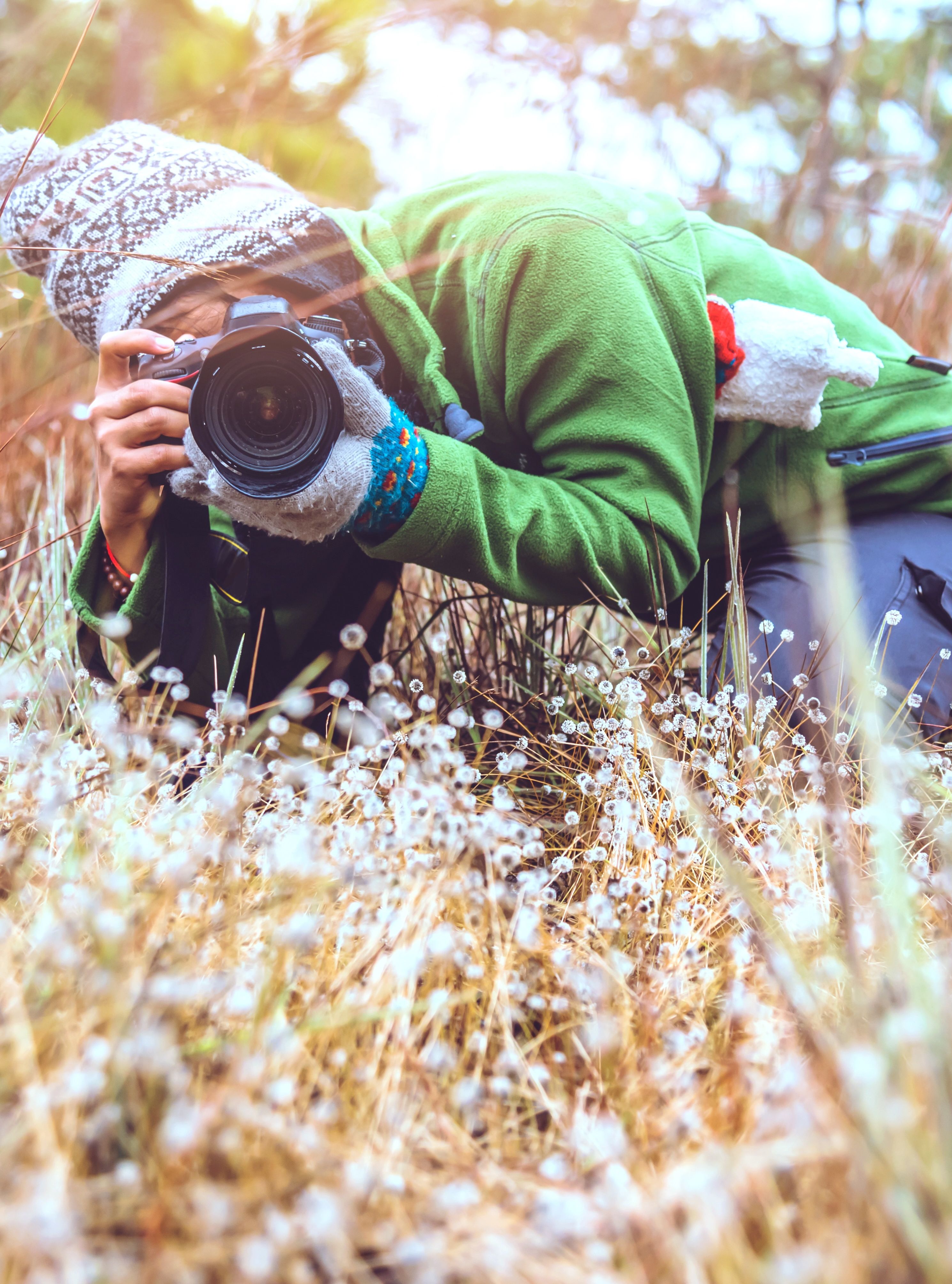 A photographer crouched low in a grassy field, aiming their camera to capture close-up shots of nature