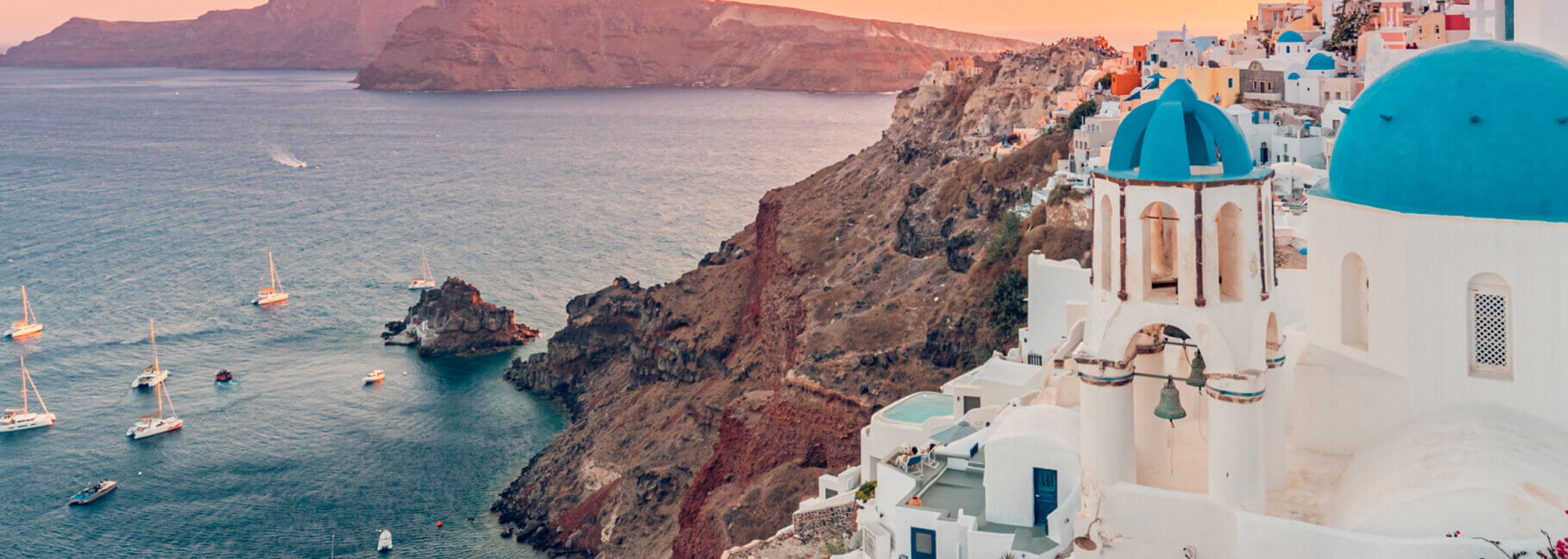 Una impresionante vista costera con edificios encalados y un acantilado escarpado con vista a un mar tranquilo salpicado de veleros durante una cálida puesta de sol.