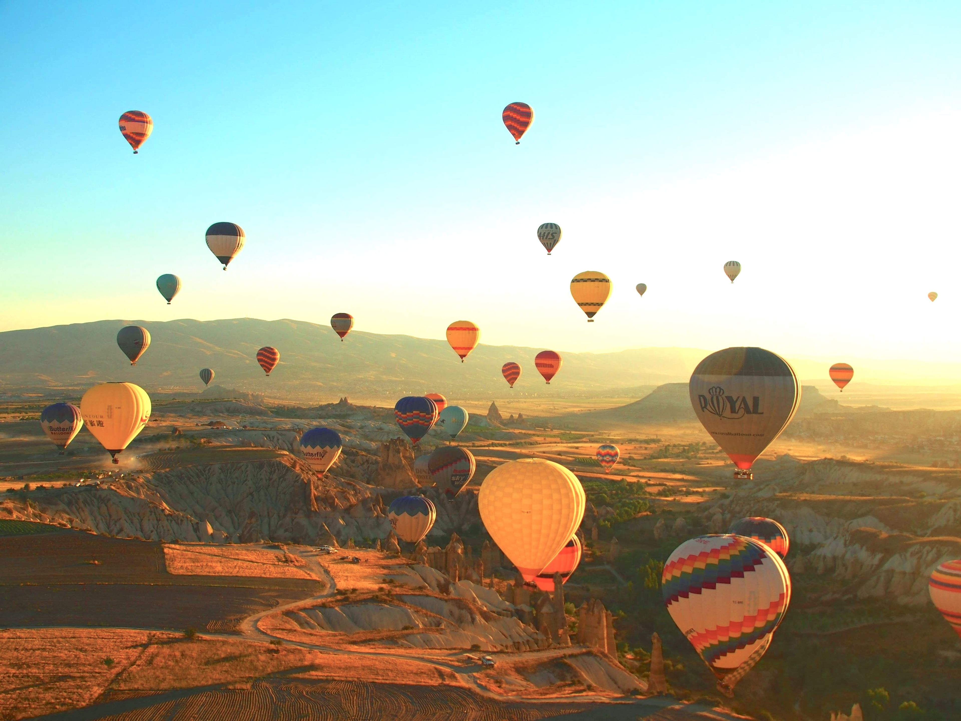 Colorful hot air balloons drifting over dramatic landscapes at sunrise, with soft golden light illuminating unique rock formations and the expansive terrain.