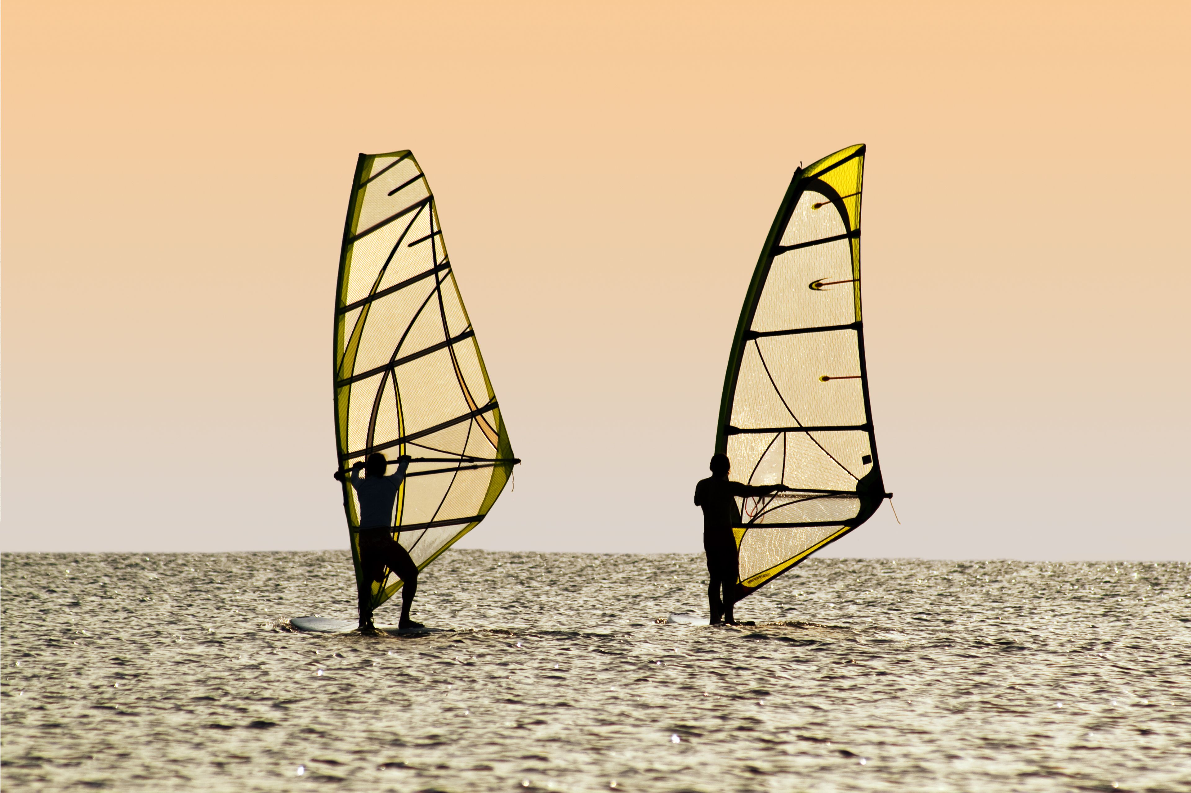 Two windsurfers on the water during sunset, balancing on their boards with their sails catching the wind