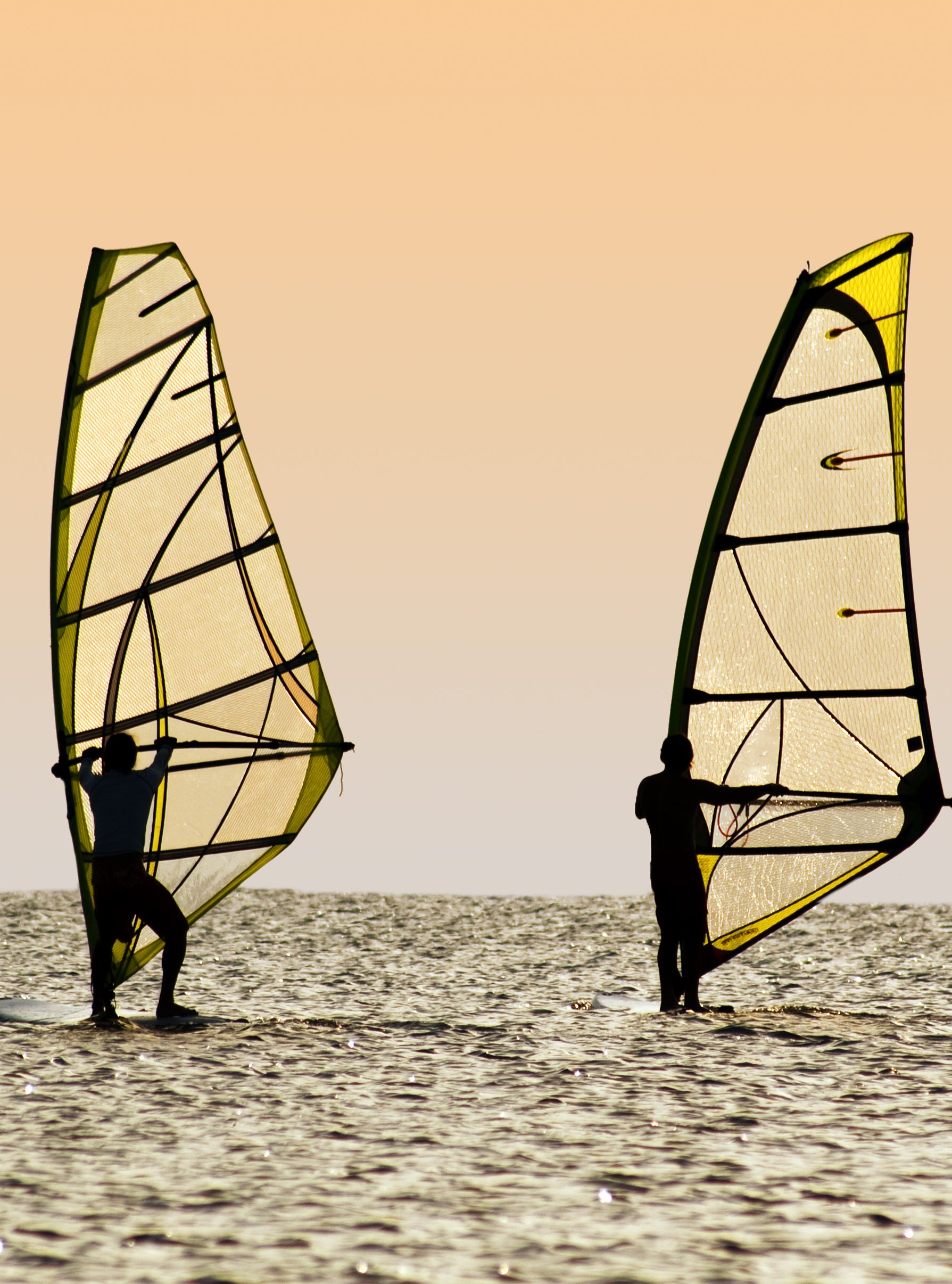 Two windsurfers on the water during sunset, balancing on their boards with their sails catching the wind