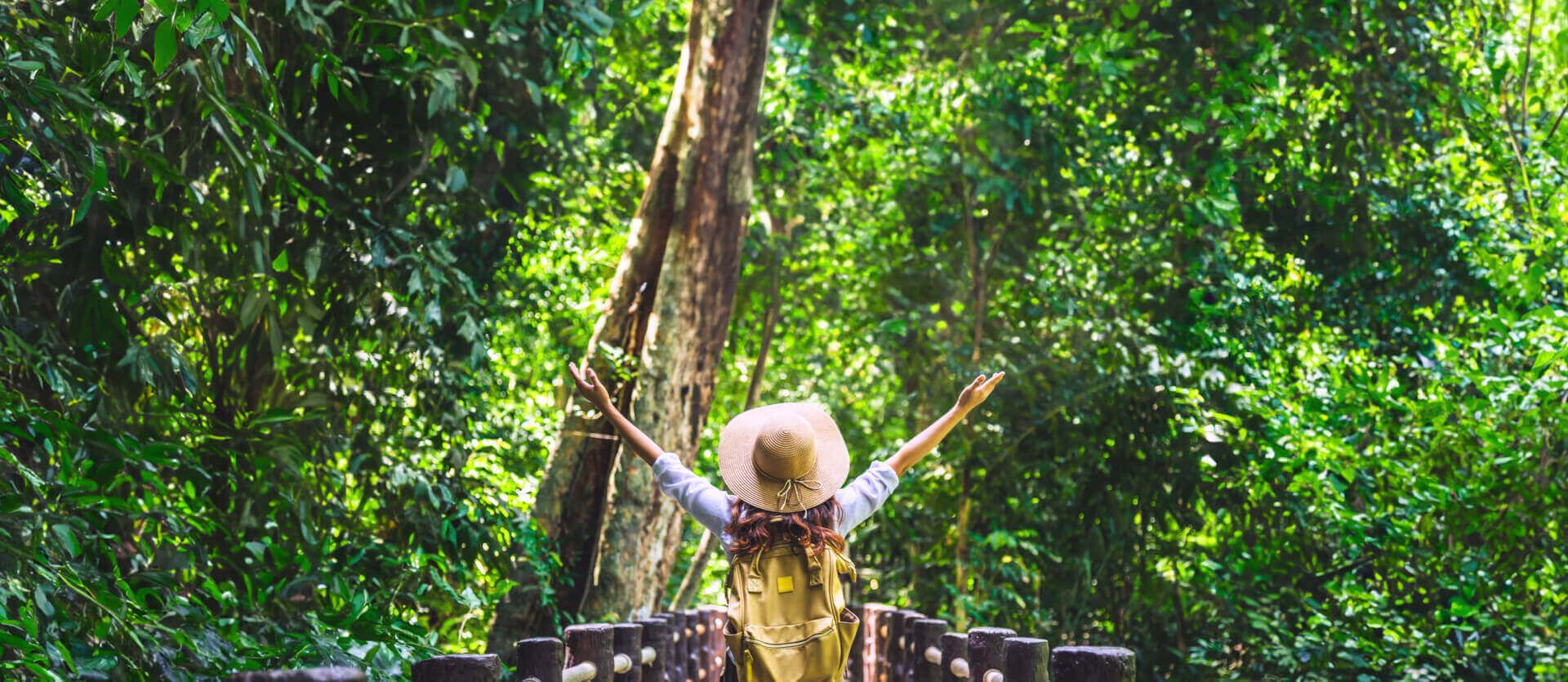 A woman with arms raised in celebration, wearing a backpack and hat, standing on a wooden bridge surrounded by lush, vibrant green forest.