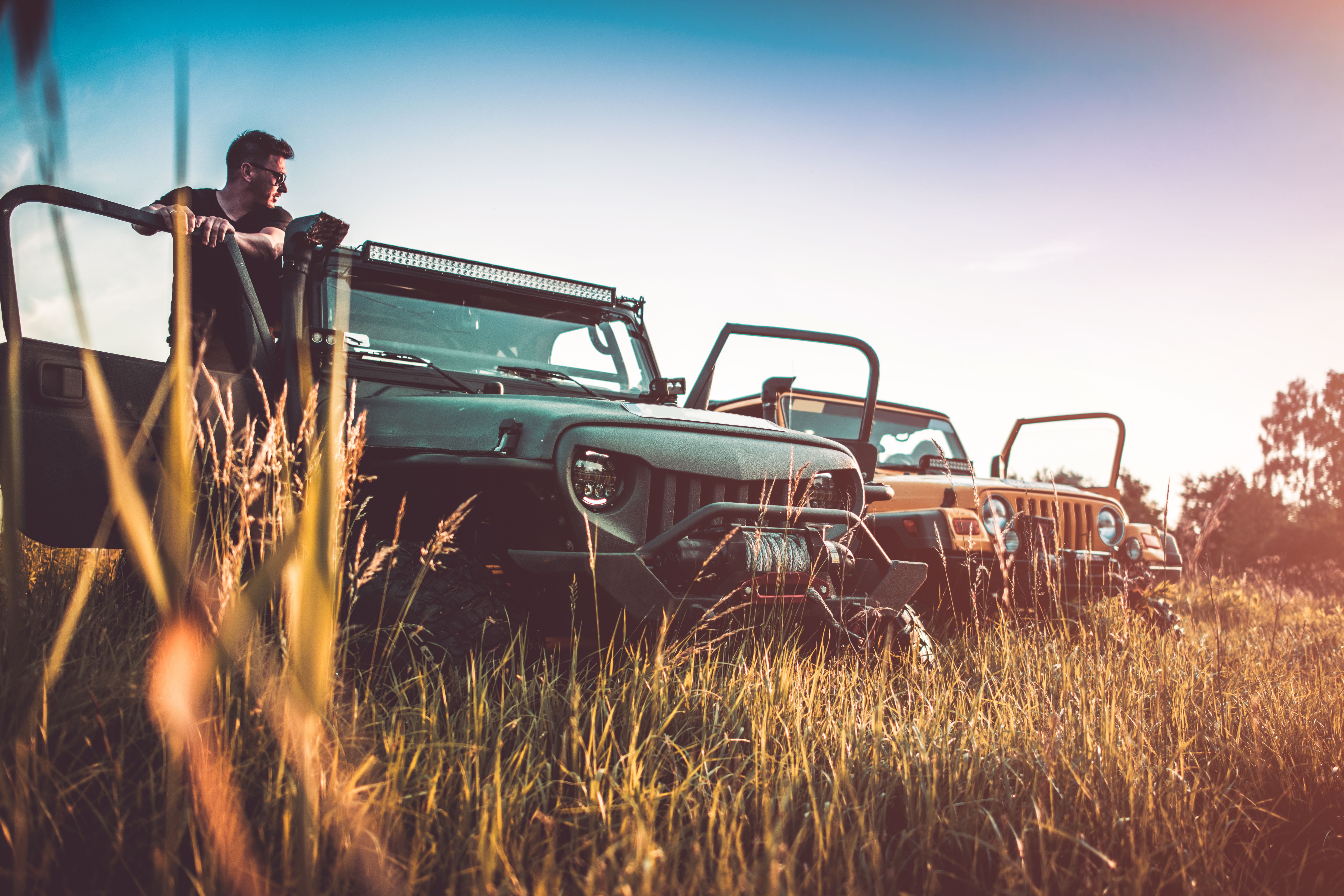Man leaning against an off-road vehicle parked in a field with two other jeeps in the background, during golden hour