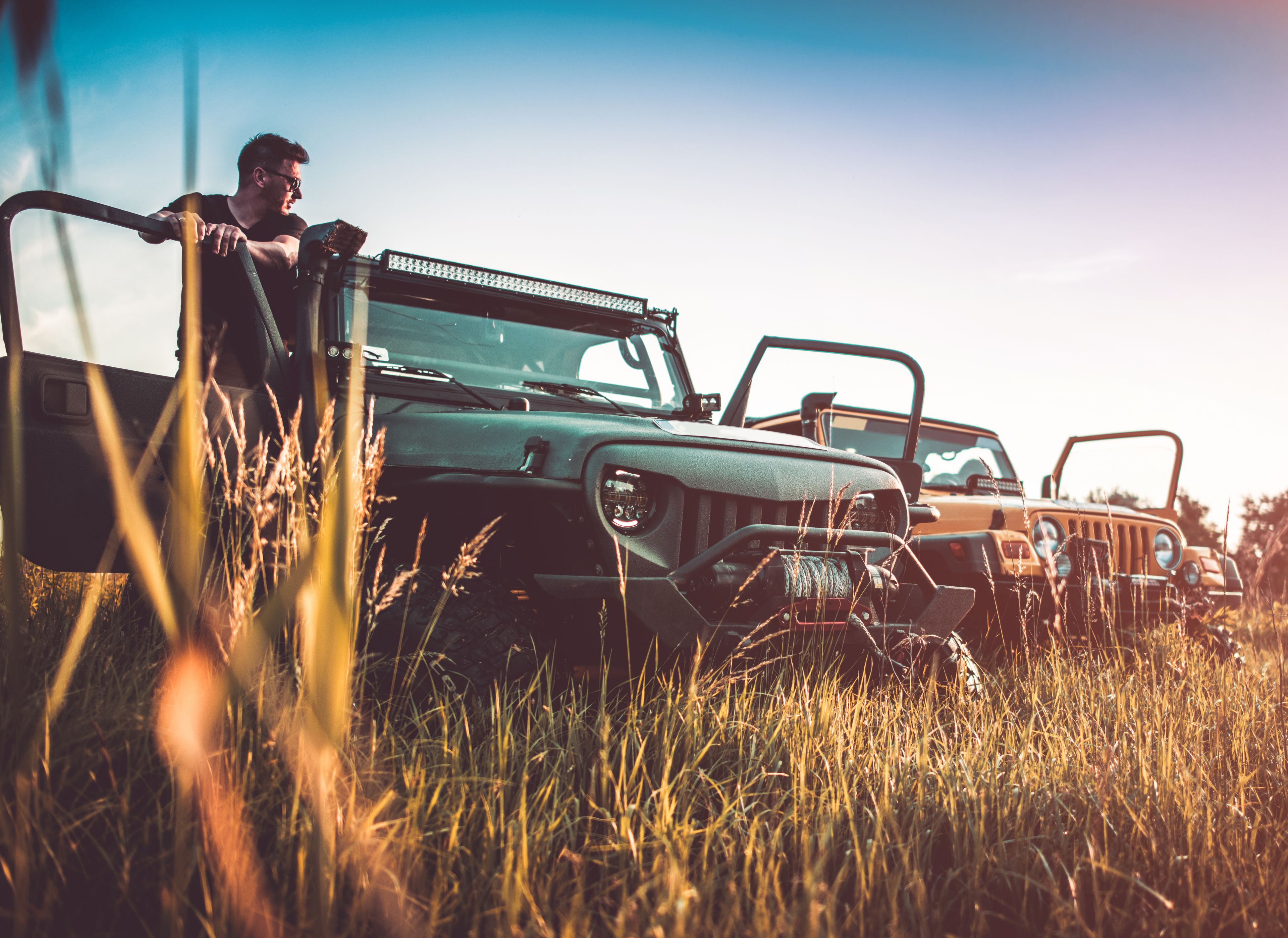 Man leaning against an off-road vehicle parked in a field with two other jeeps in the background, during golden hour