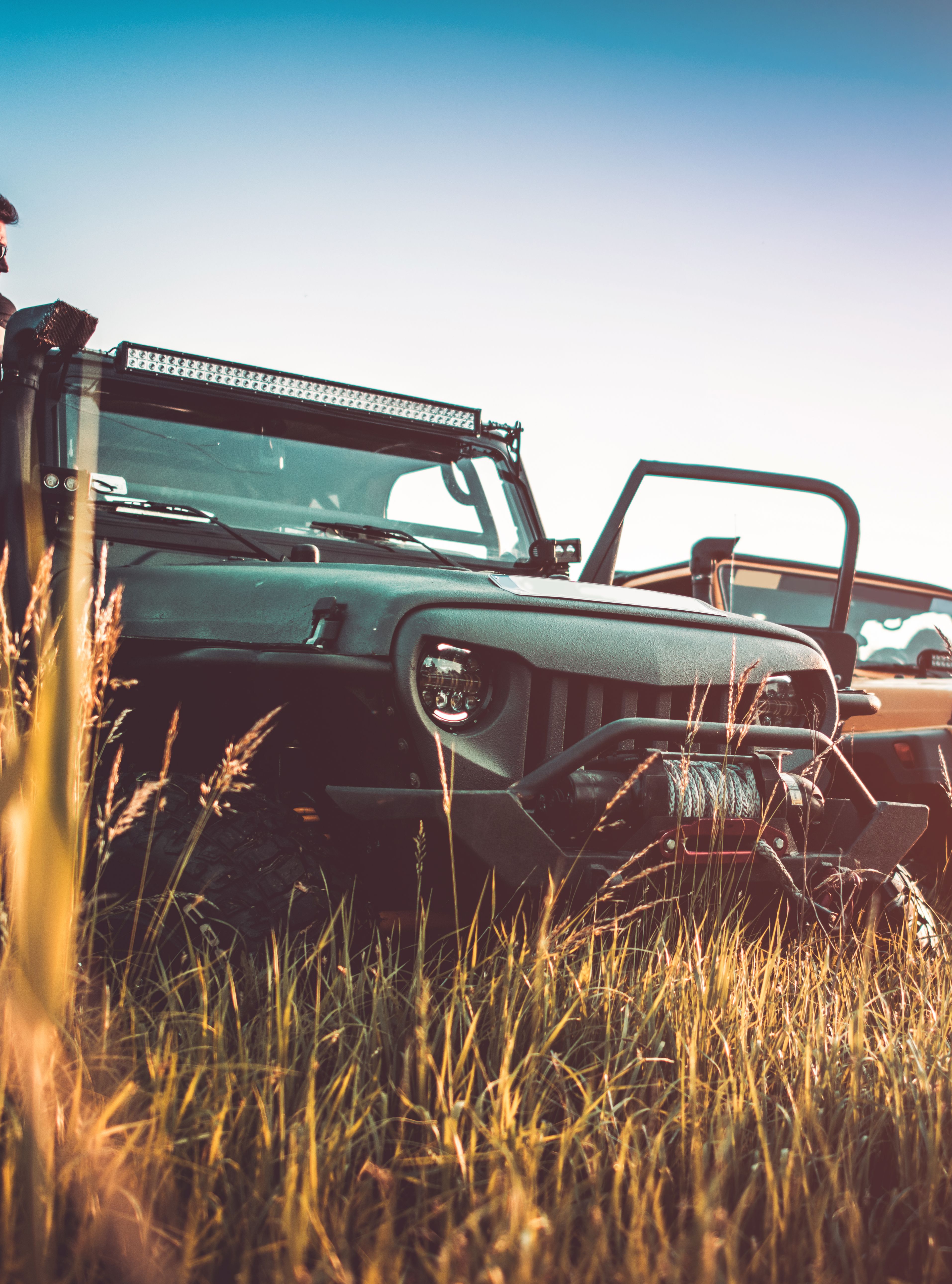 Man leaning against an off-road vehicle parked in a field with two other jeeps in the background, during golden hour