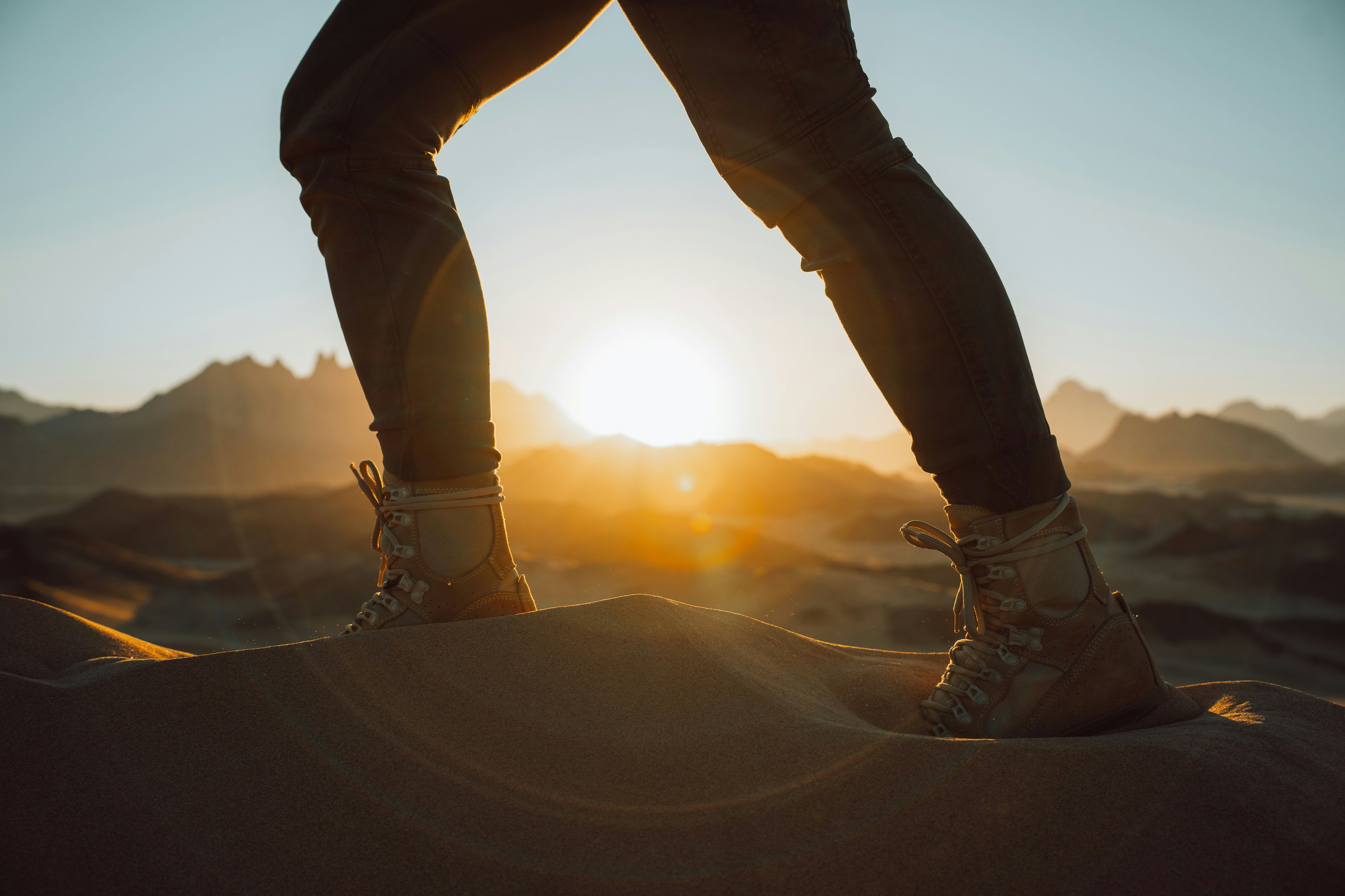 Person standing on a sand dune, with hiking boots, overlooking the sun setting behind distant mountains