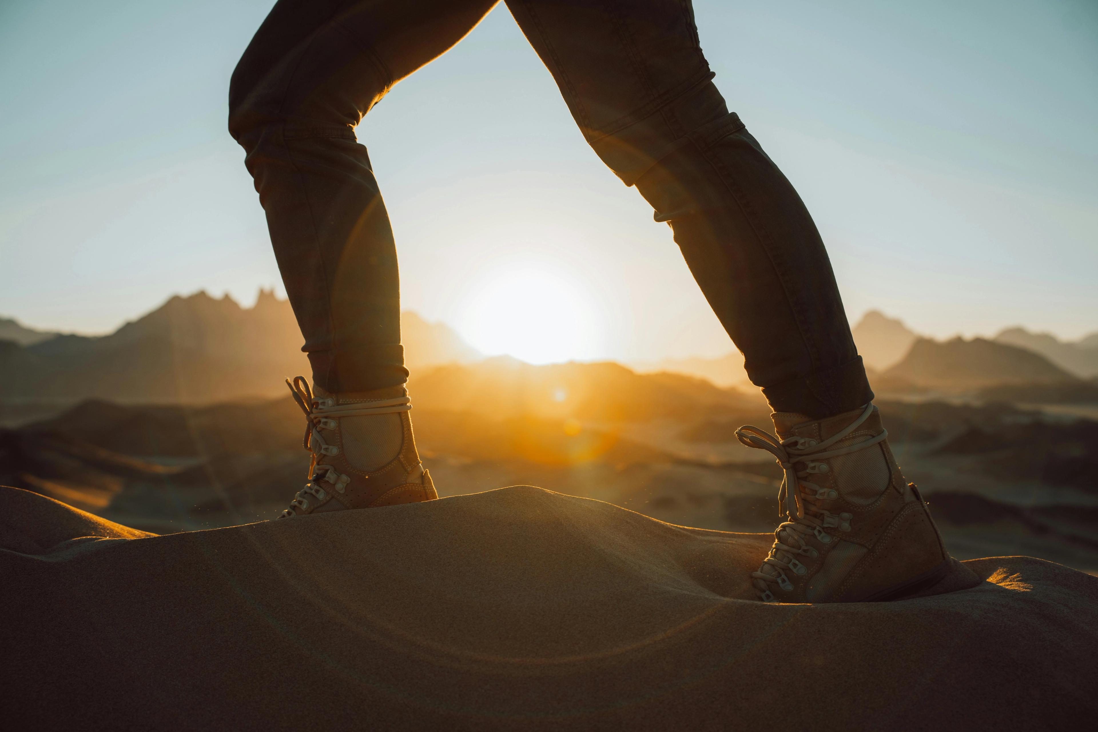 Person standing on a sand dune, with hiking boots, overlooking the sun setting behind distant mountains