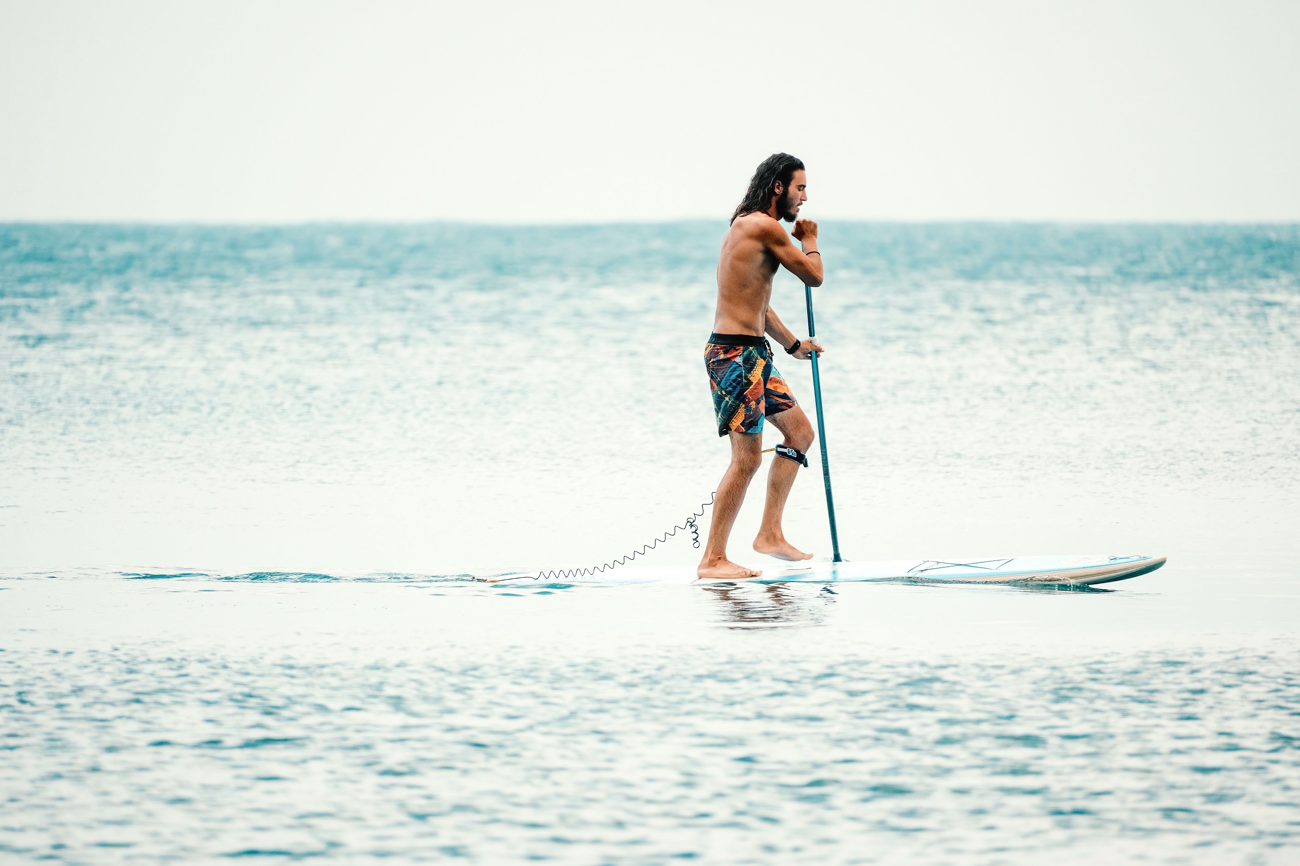 Man stand-up paddleboarding on calm water, wearing patterned shorts and navigating with a paddle