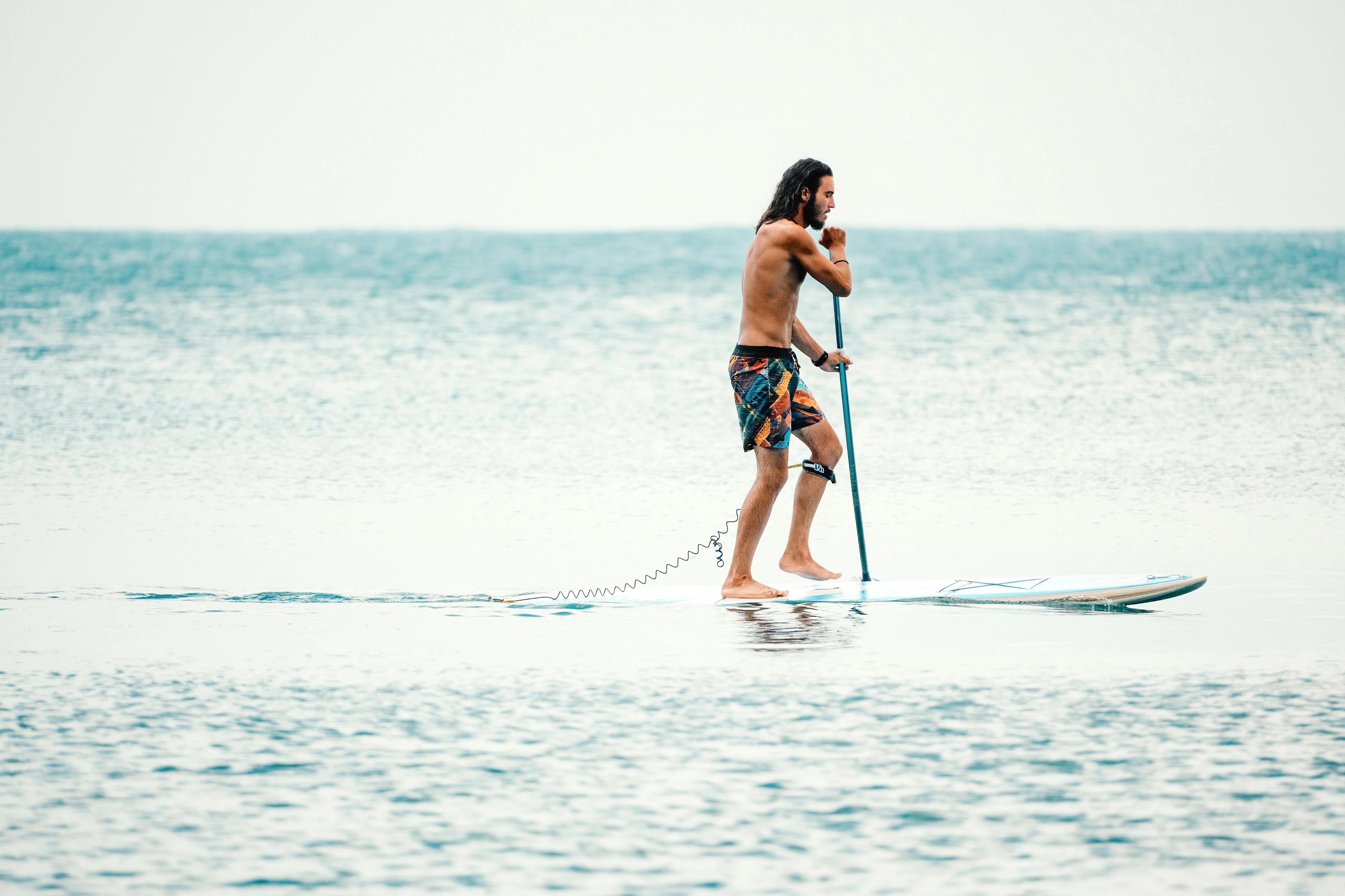 Man stand-up paddleboarding on calm water, wearing patterned shorts and navigating with a paddle