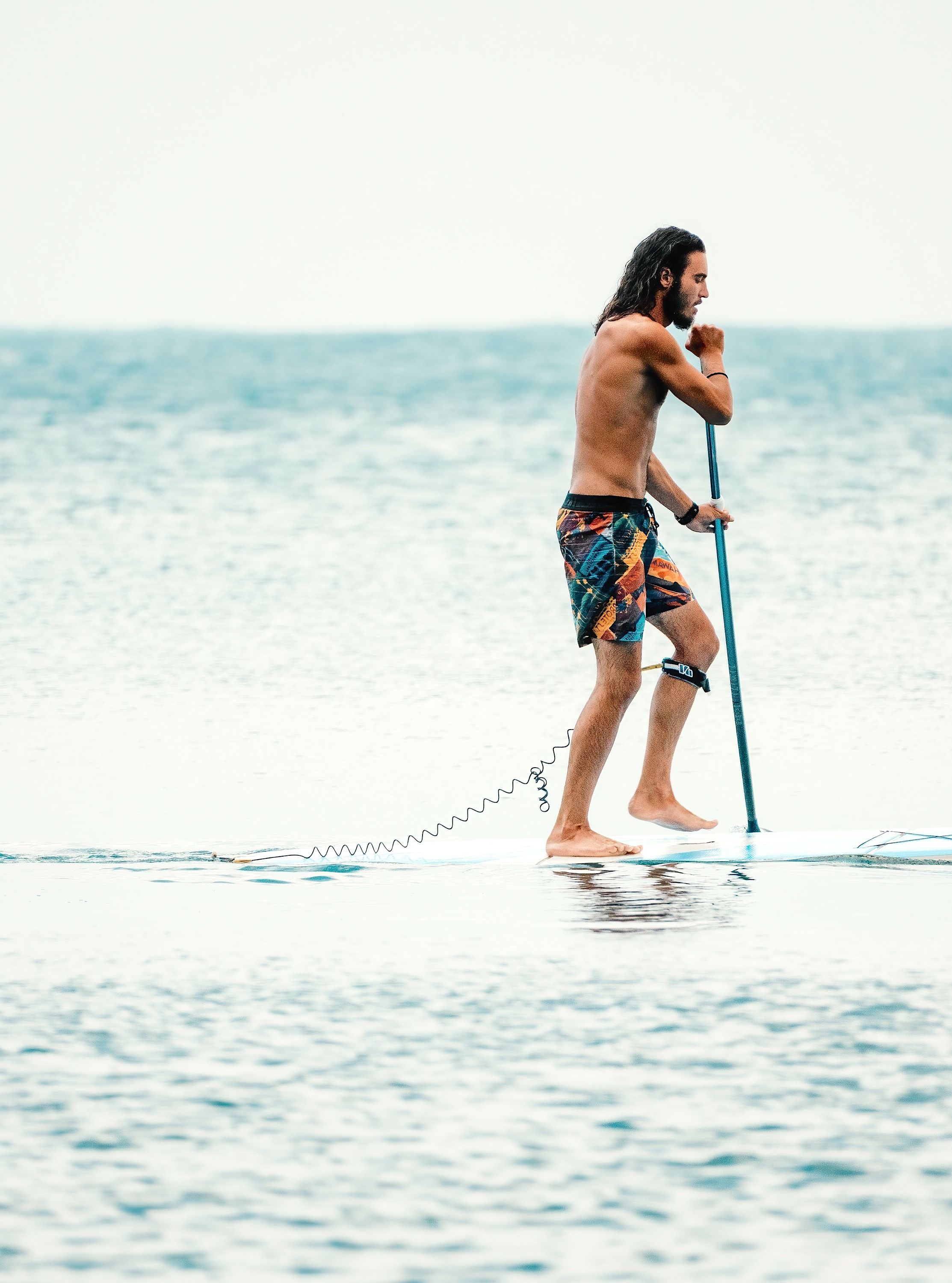 Man stand-up paddleboarding on calm water, wearing patterned shorts and navigating with a paddle