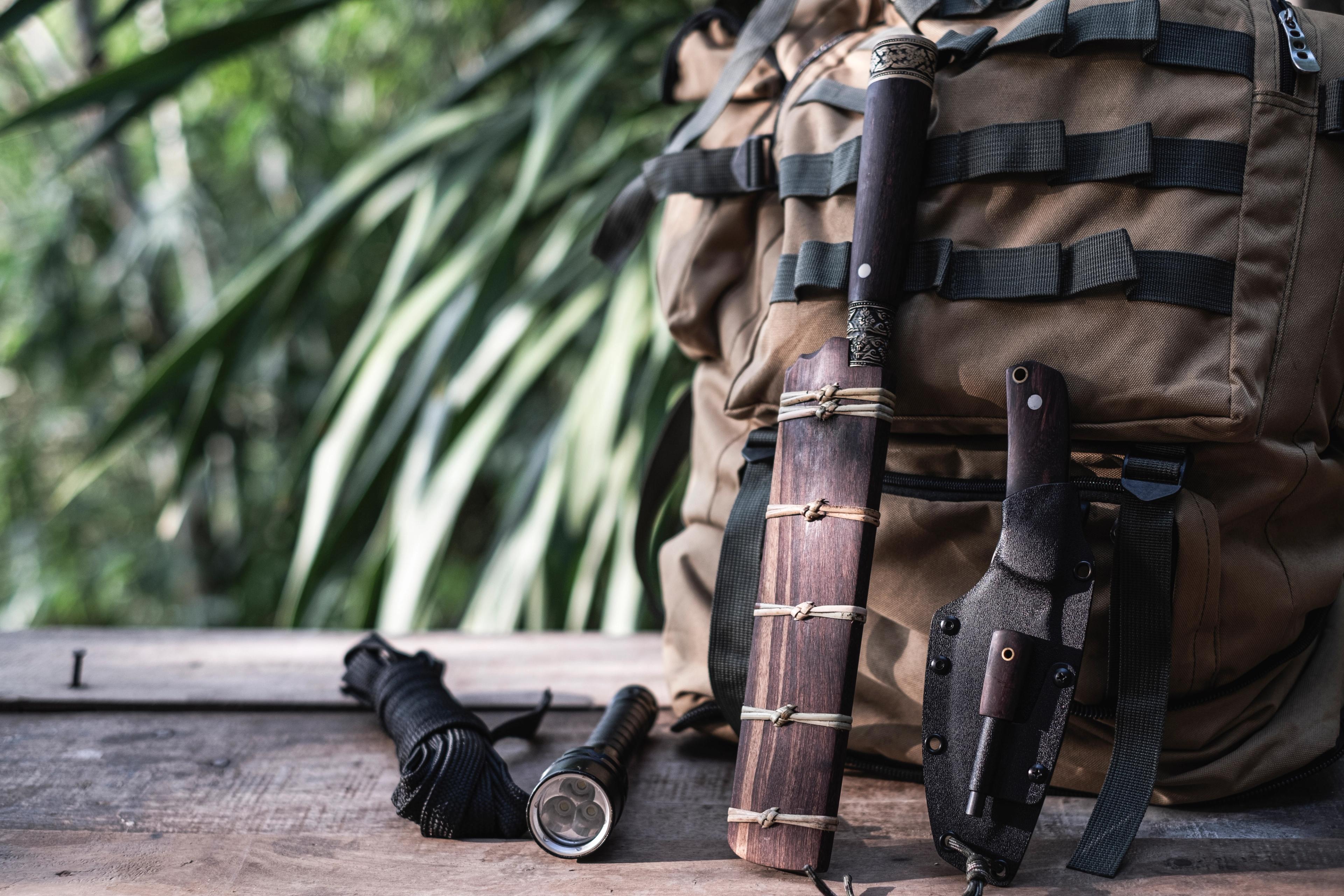 Survival gear including a backpack, wooden knife, flashlight, and coiled rope laid out on a wooden surface