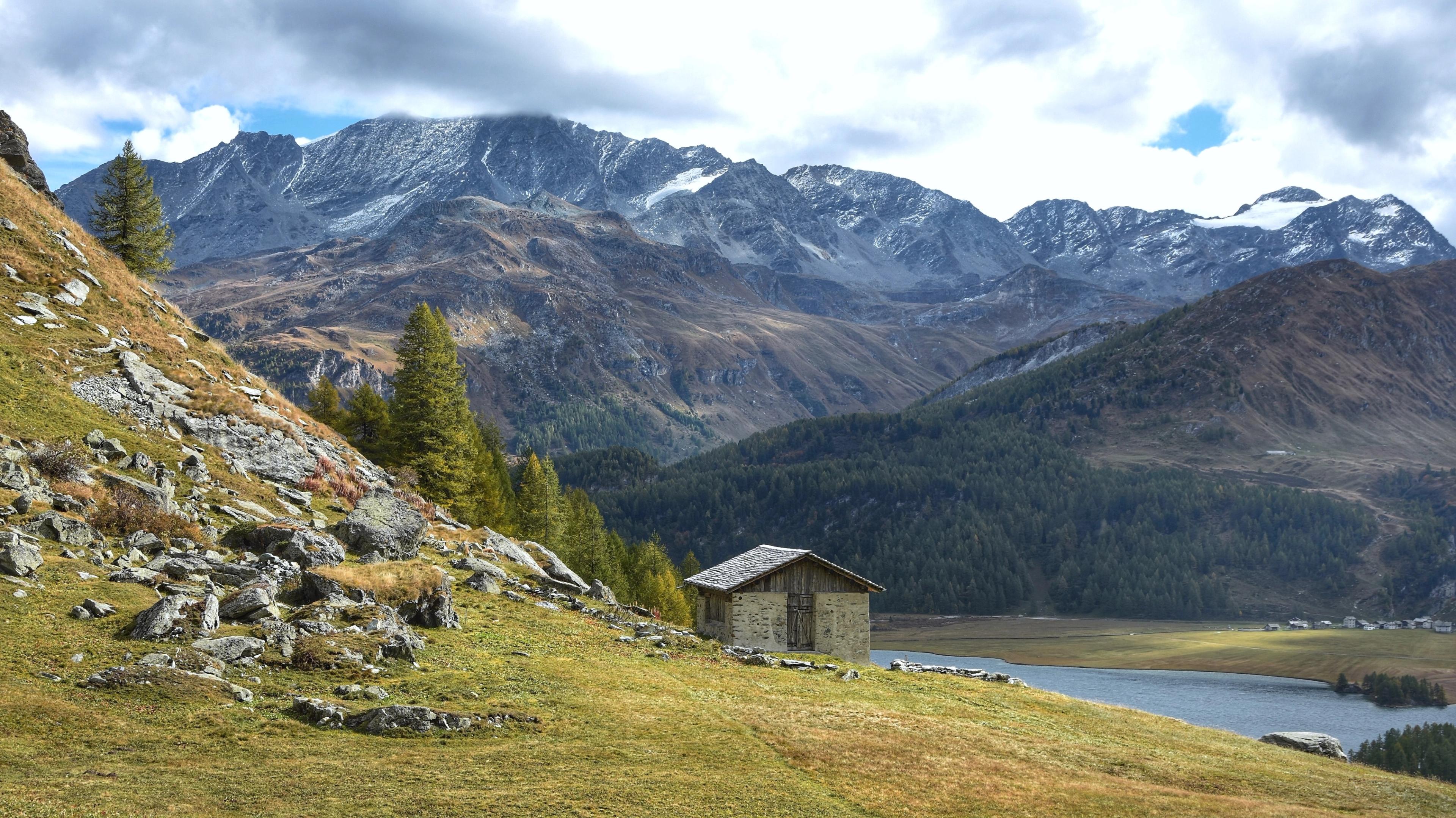 Small stone cabin nestled in a mountainous valley, with a scenic view of snow-capped peaks and lush forests