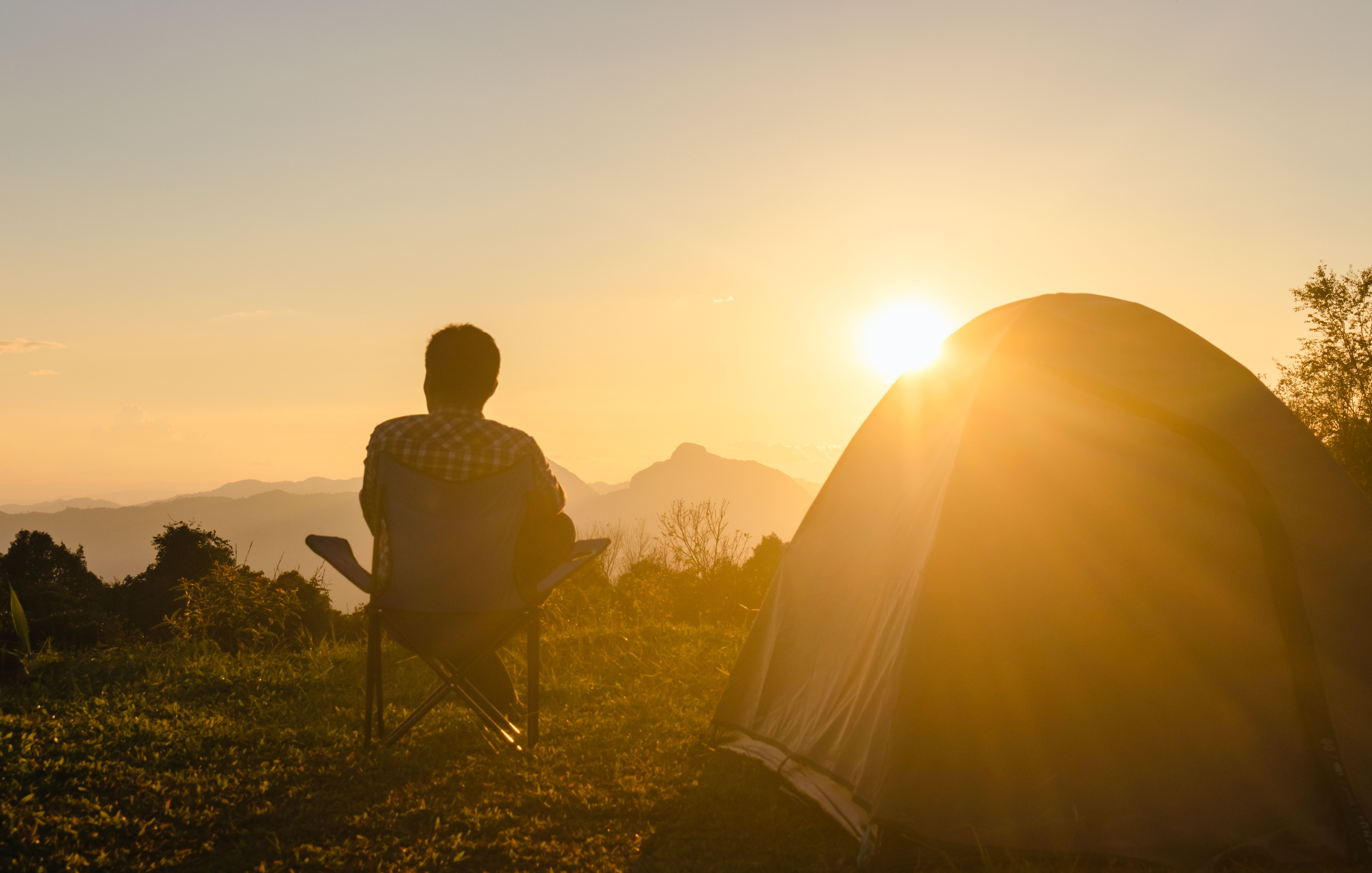 Person sitting in a camping chair beside a tent, watching the sunrise over a mountain range