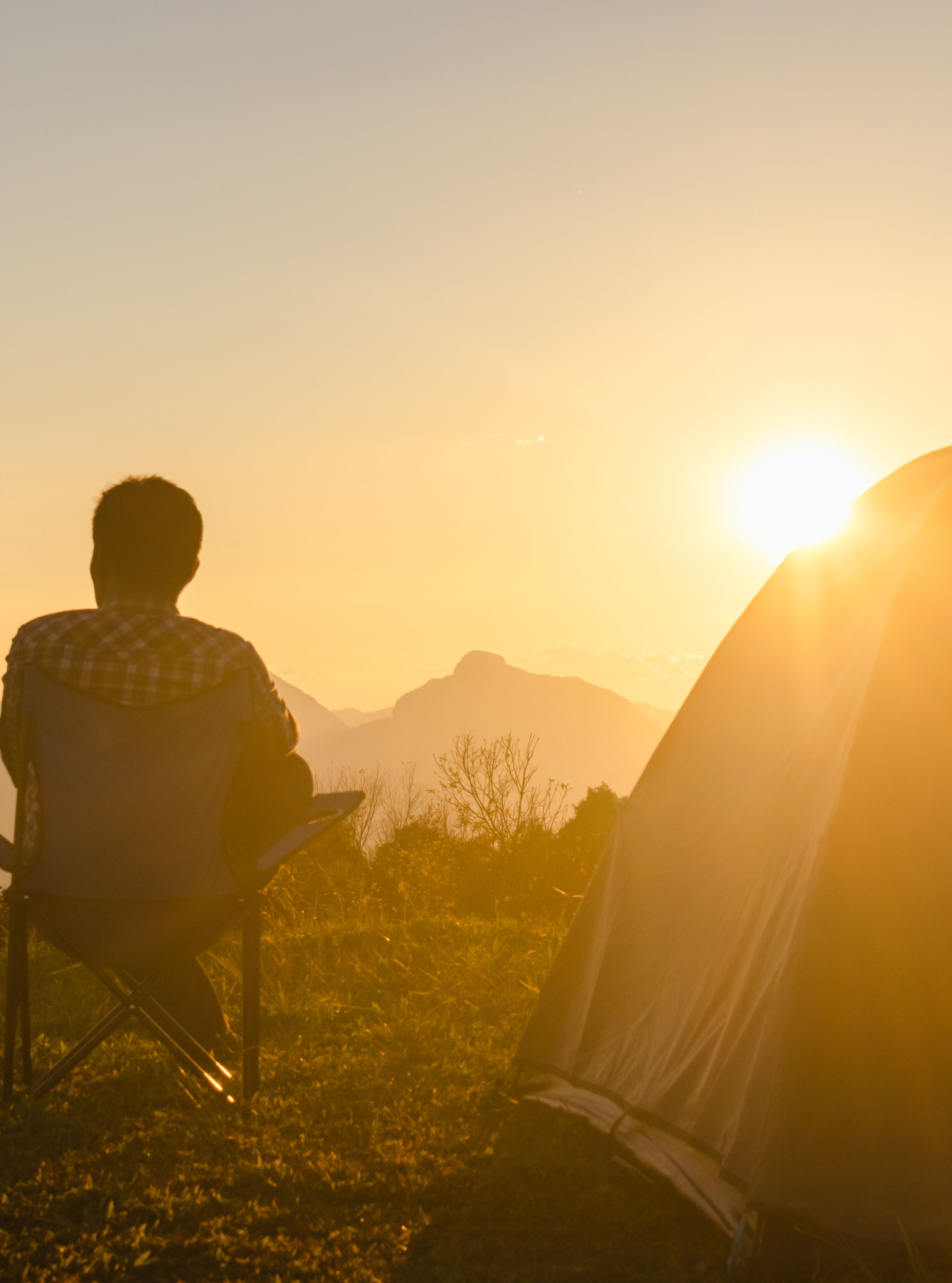 Person sitting in a camping chair beside a tent, watching the sunrise over a mountain range