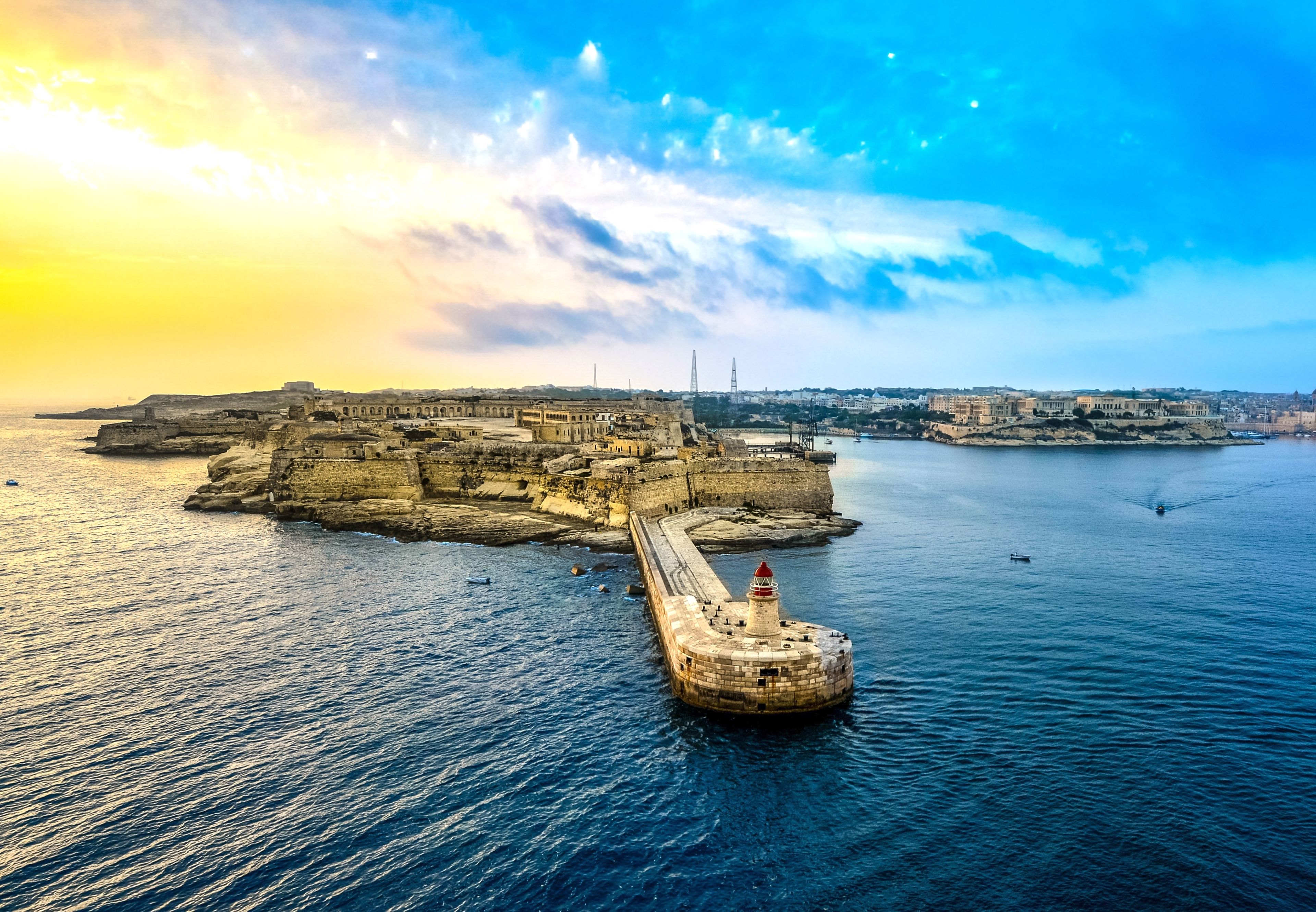 A stunning view of Fort Ricasoli in Malta at sunrise, with the vibrant yellow and blue hues of the sky reflecting on the calm sea, and the historic lighthouse marking the entrance to the Grand Harbour