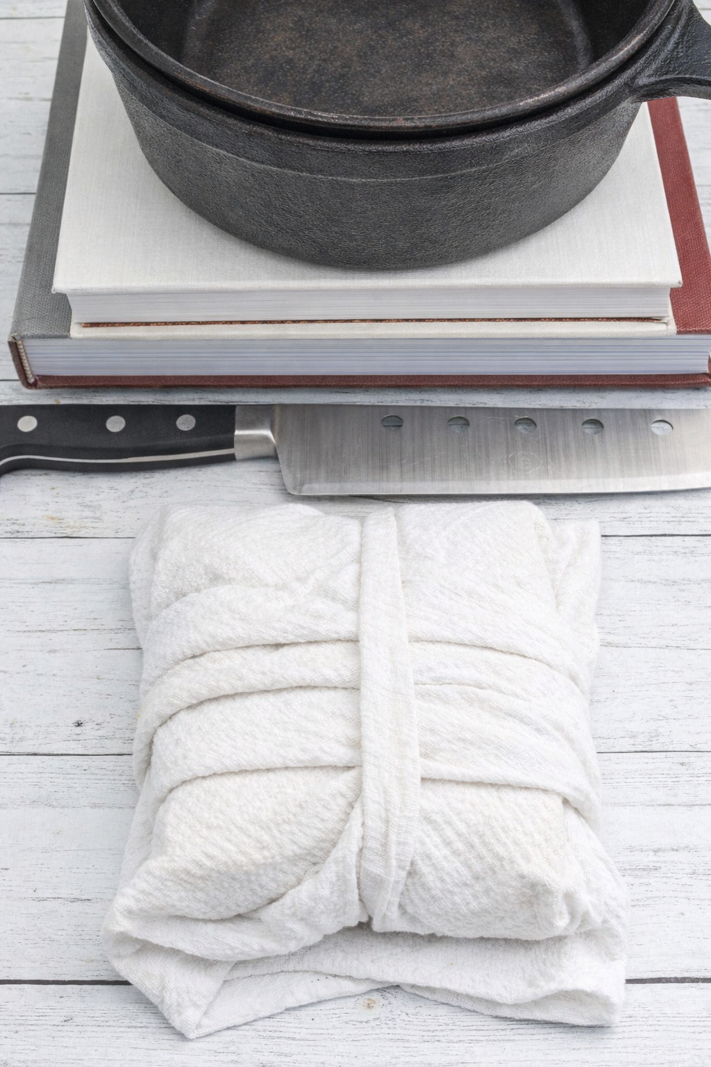 Pressed extra-firm tofu block wrapped in white kitchen towel with cast iron skillet, knife, and cutting board on wooden surface for water removal.