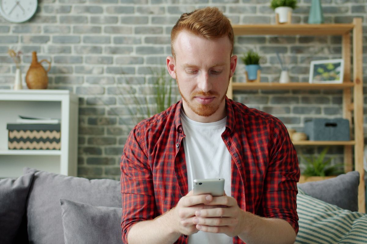 Young man with red hair in a checked shirt looking down at his smartphone while sitting on a grey sofa at home
