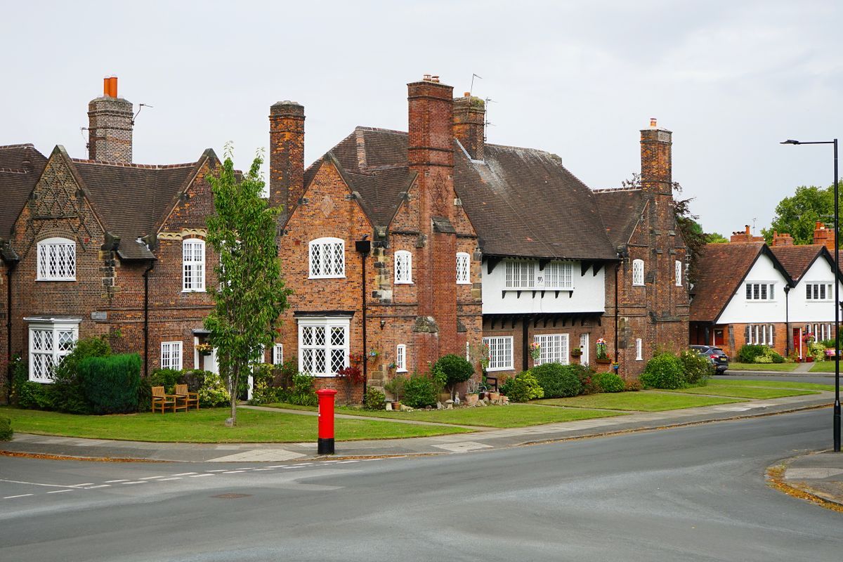 Red brick houses with tall chimneys and a white-rendered upper floor behind a lawn, with a red postbox on a quiet street corner