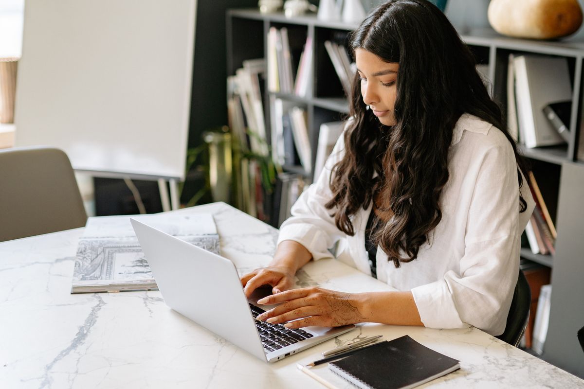 Woman in a white shirt typing on a laptop at a white marble desk with a flip chart and bookshelves behind her