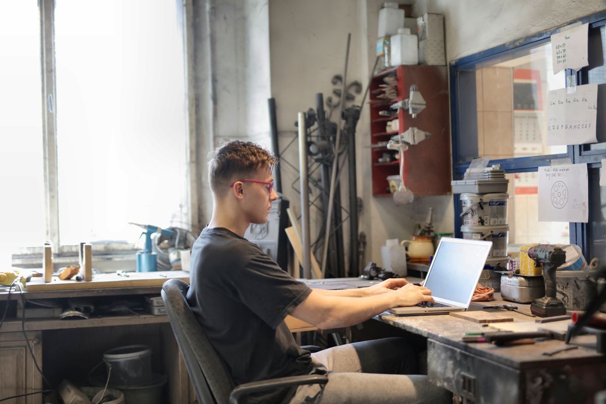 Young man in glasses typing on a laptop at a cluttered workbench in a workshop with tools on the walls