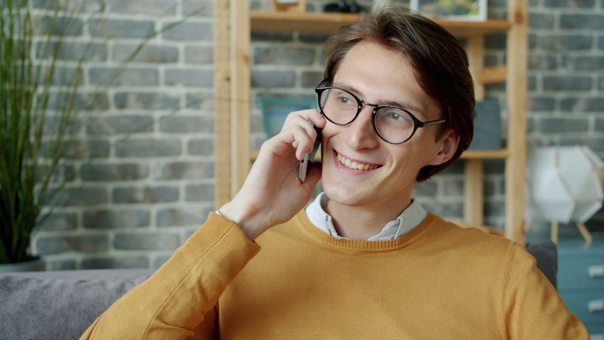 Smiling man in glasses and a mustard jumper talking on a mobile phone at home