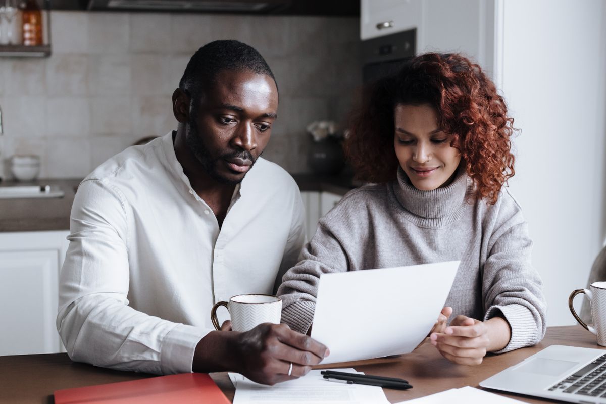 Man and woman at a kitchen table reading a printed letter together, with mugs, pens and an open laptop in front of them