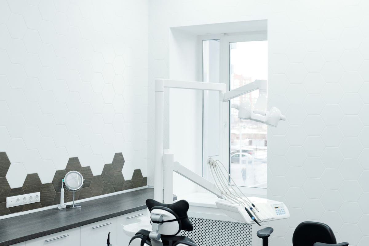 Bright dental surgery with a white dental unit and overhead light, a black treatment chair and a dark worktop beside a window