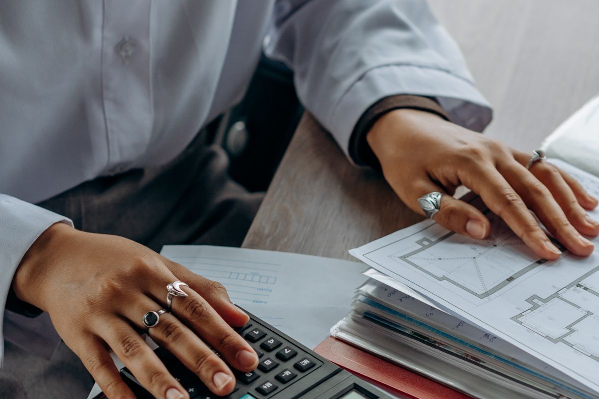 Hands with silver rings using a calculator beside a stack of folders and a printed floor plan on a desk