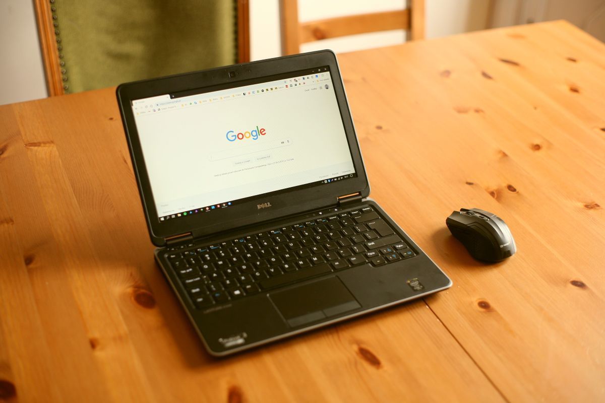 Black laptop on a wooden table showing the Google search page, with a wireless mouse beside it