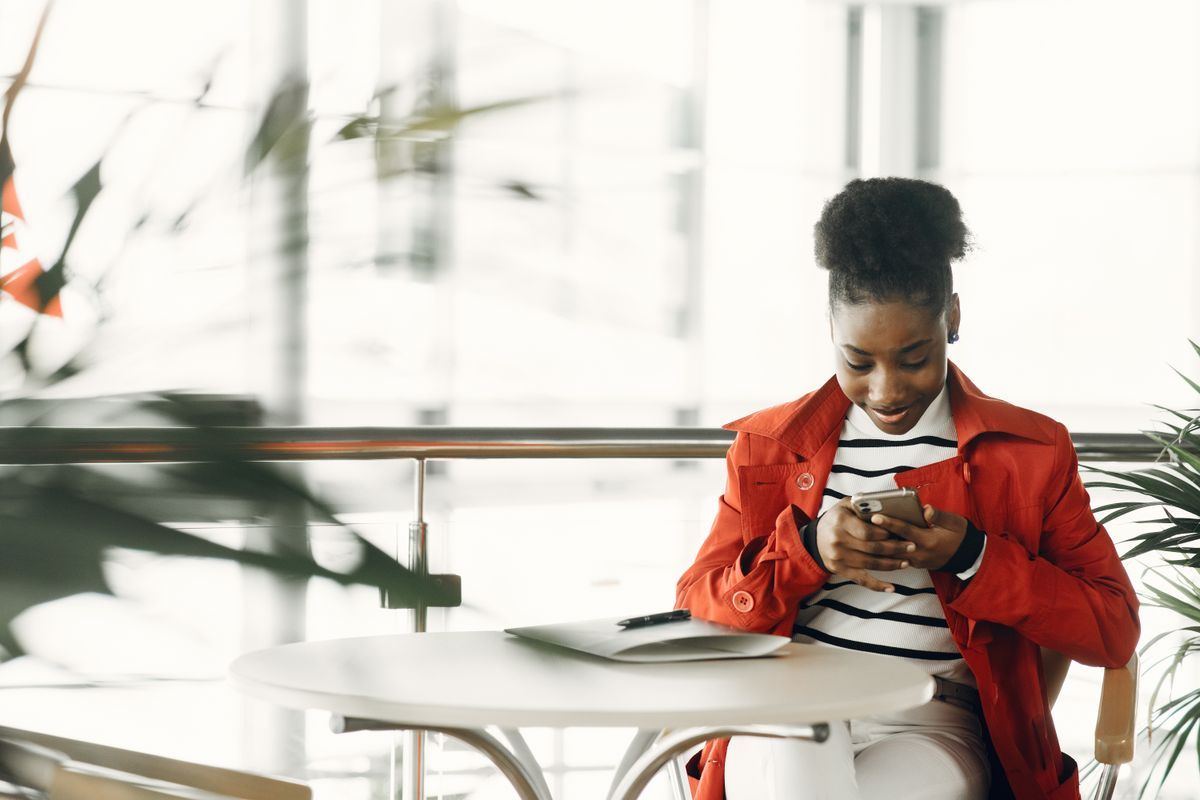 Young woman in a red jacket looking at her smartphone while sitting at a round white table in a bright café