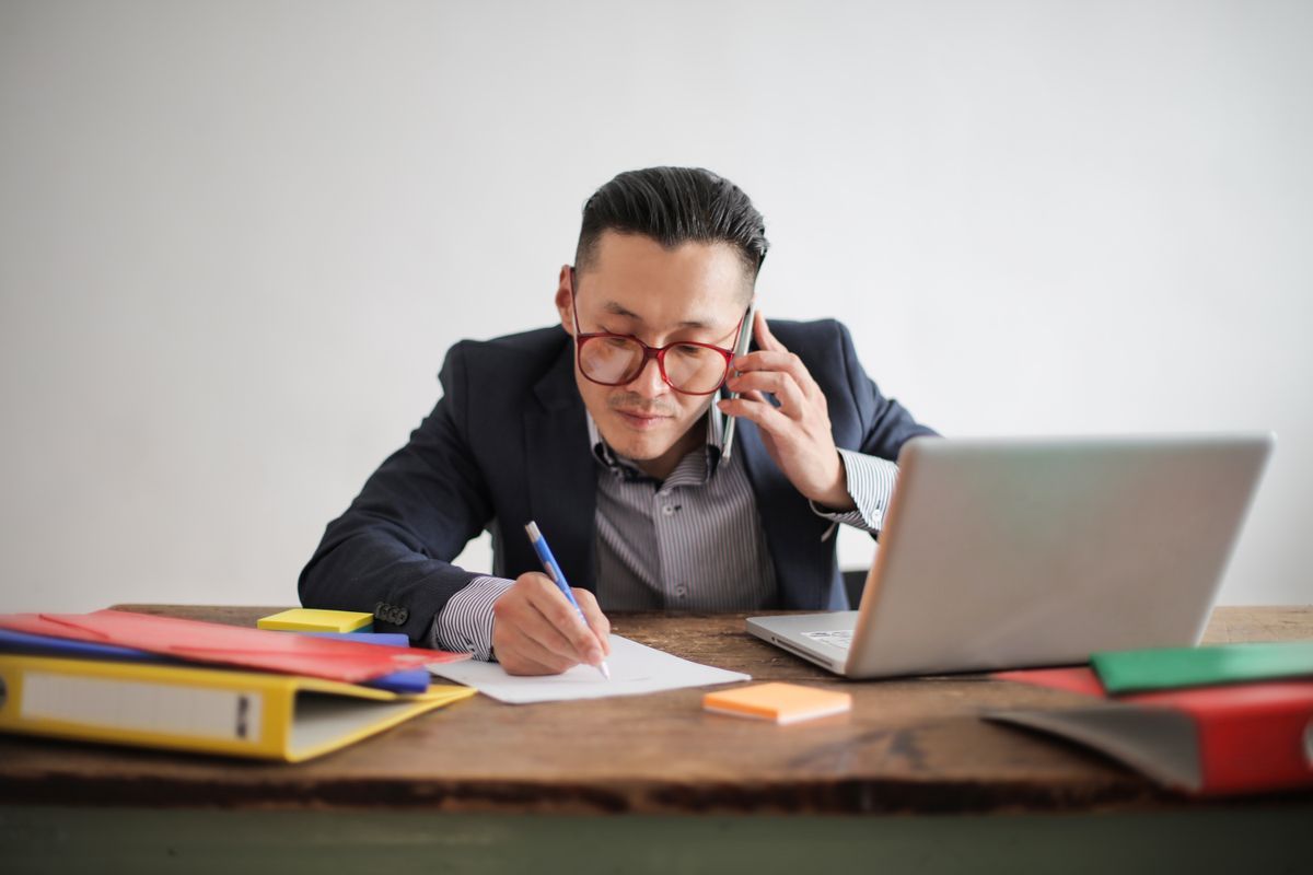 Man in a navy jacket and glasses talking on a mobile phone and writing notes at a desk beside an open laptop and coloured folders