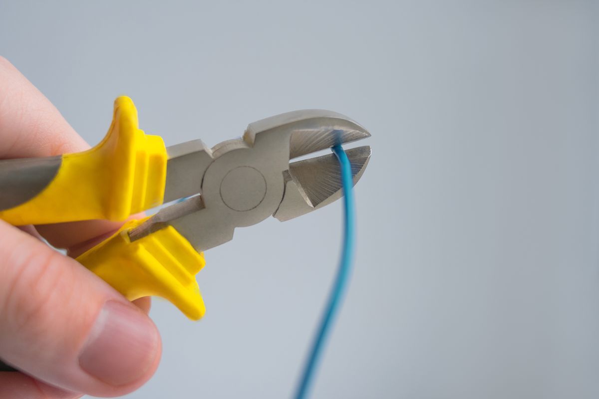 Hand holding yellow-handled side cutters to snip a blue electrical wire against a plain grey background