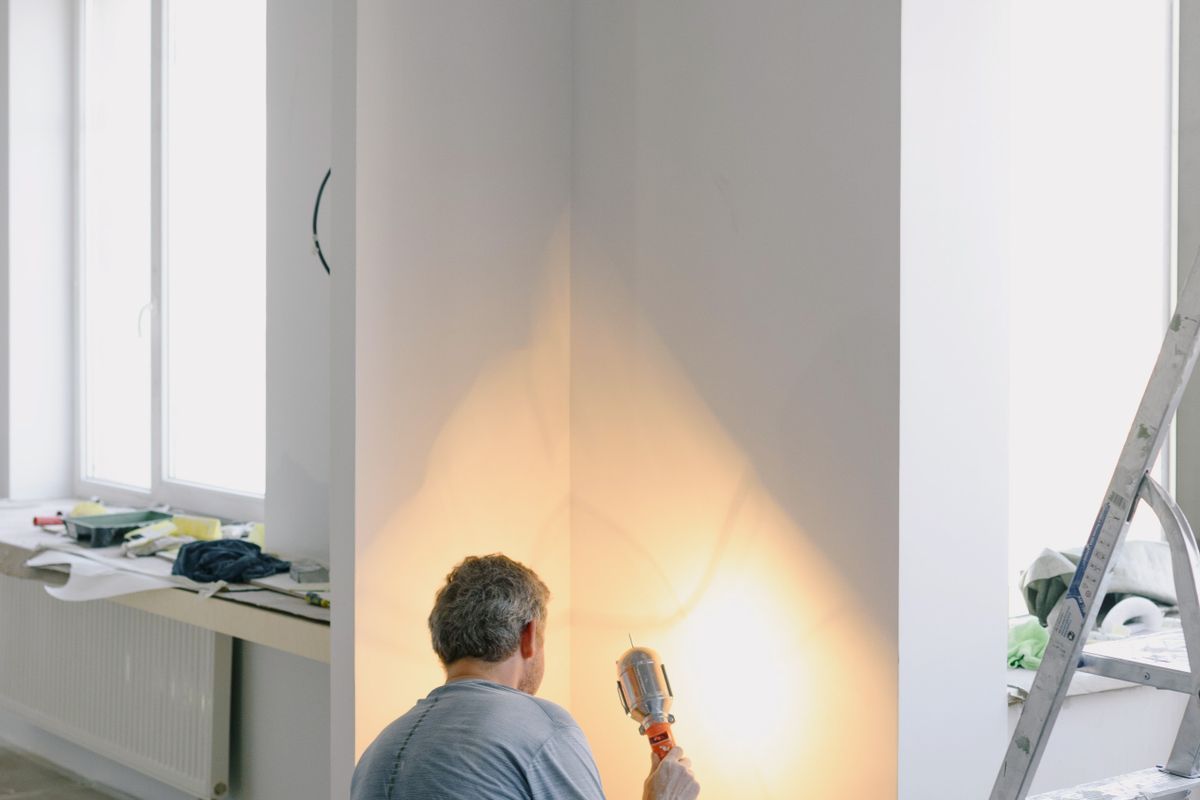 Grey-haired tradesman crouching in a freshly painted white room, holding a work lamp up to the wall, with a stepladder beside him