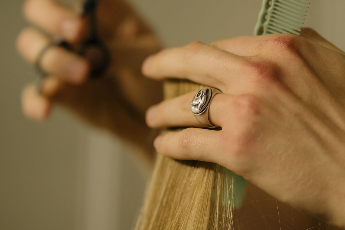 Stylist's hand with a silver ring holding a section of long blonde hair and a mint green comb, with scissors blurred in the background