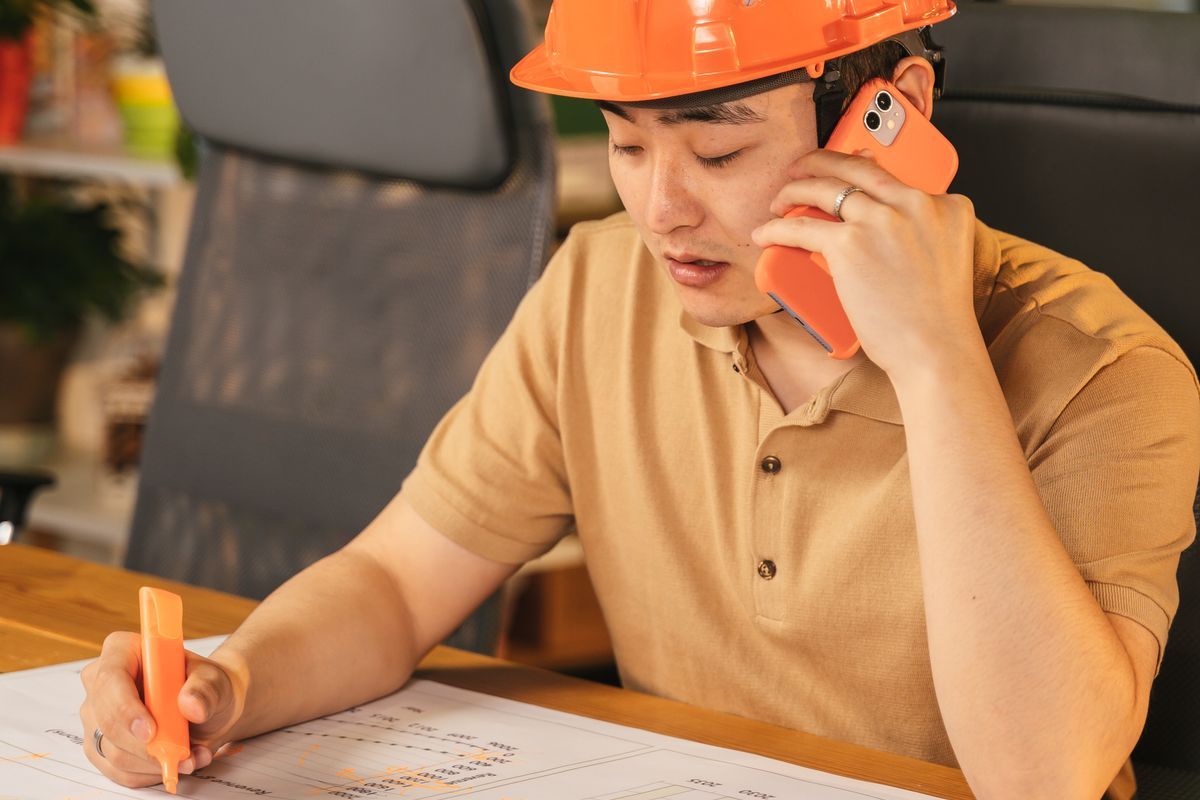 Man in an orange hard hat talking on an orange mobile phone while marking a paper plan with a highlighter at a wooden desk
