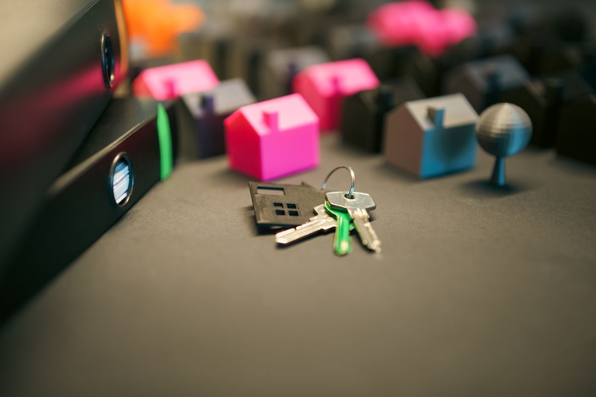 Keys on a house-shaped key ring in front of small model houses, beside black lever-arch files on a grey desk