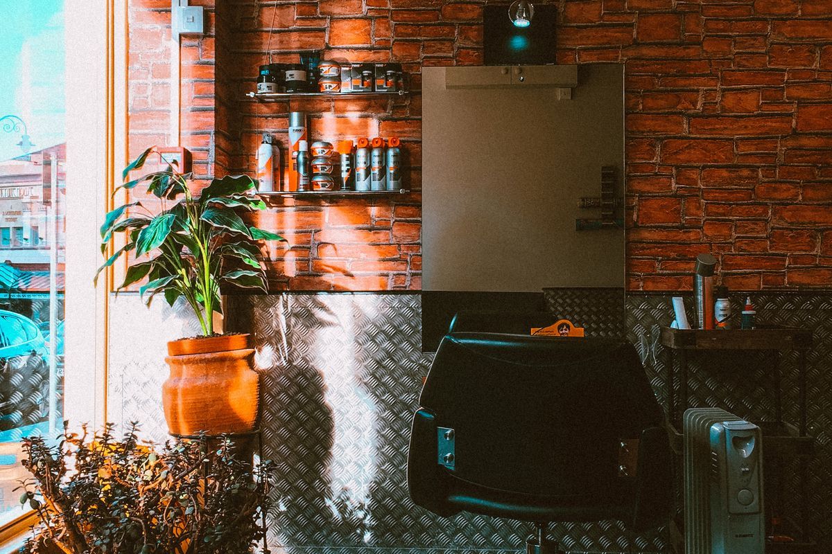 Barber chair in front of a mirror on an exposed brick wall, with product shelves, a potted plant by the window and a small oil heater