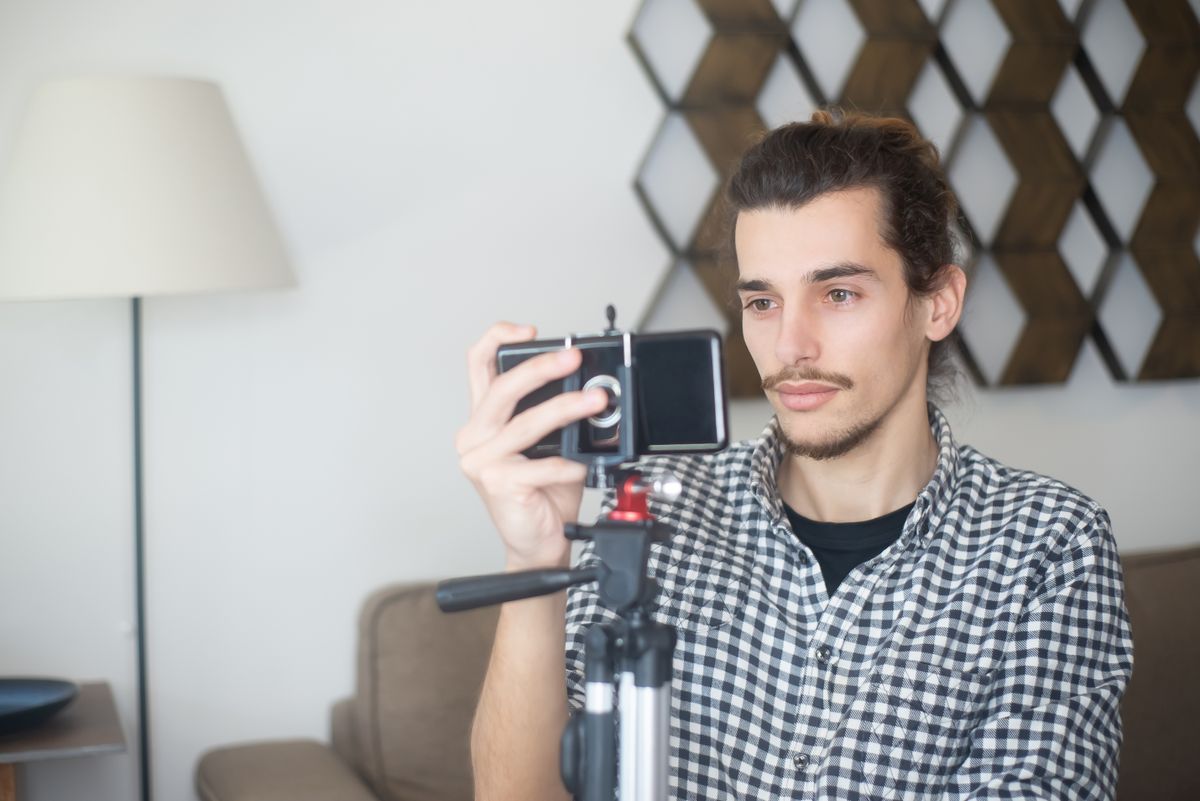 Young man in a checked shirt framing a shot on a smartphone mounted on a tripod in a living room