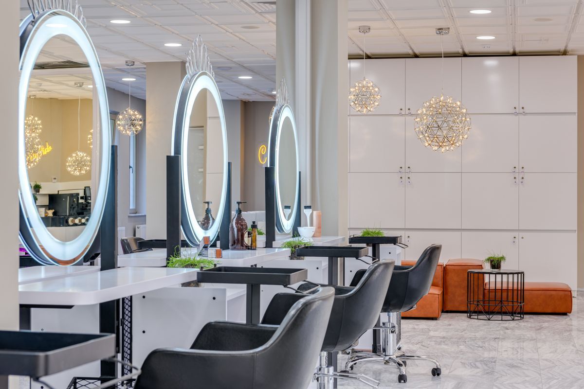 Salon floor with black styling chairs facing round illuminated mirrors, white workstations, sparkling pendant lights and tan leather seating at the back