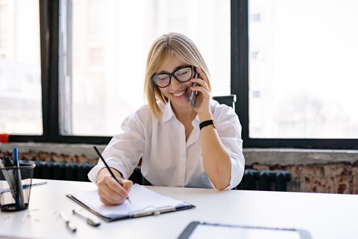 Smiling woman in glasses talking on a mobile phone and writing notes on a clipboard at a white desk by a large window