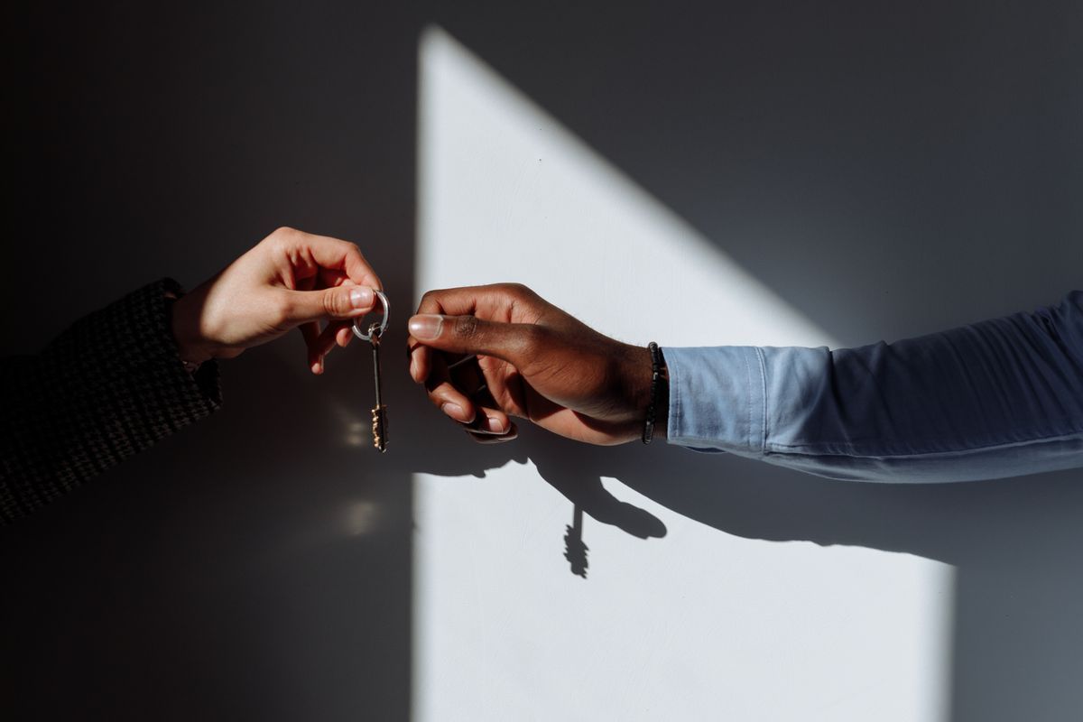 Two hands passing a single key on a ring across a sunlit white wall, one in a tweed sleeve and one in a blue shirt