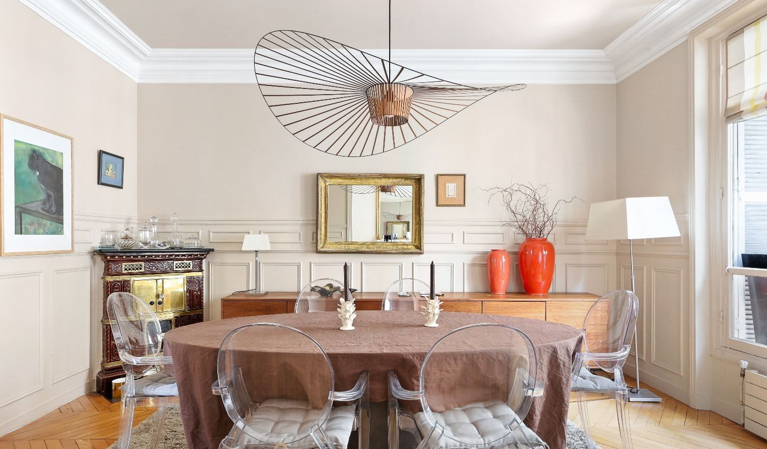 Salle à manger haussmannienne d'un appartement meublé rue Lentonnet Paris 9e, suspension Vertigo au-dessus d'une table ovale et chaises Louis Ghost sous des moulures d'époque